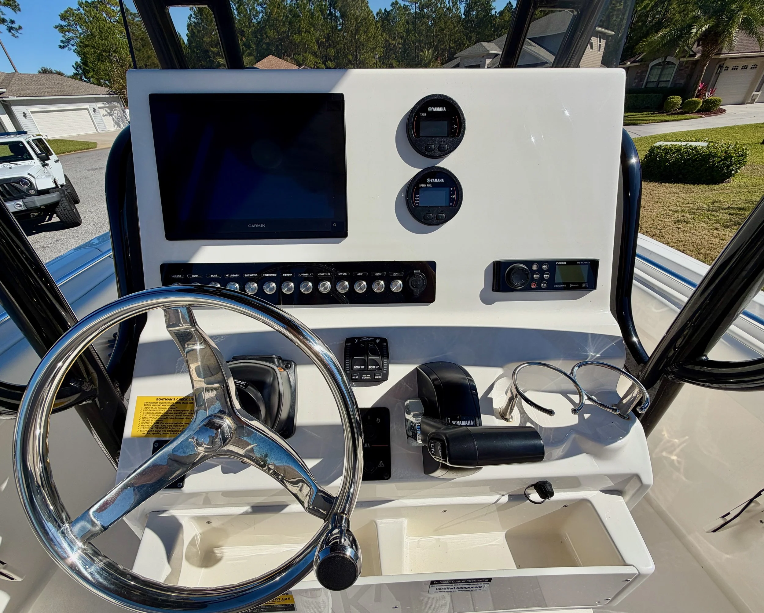 Boat cockpit with steering wheel, electronic controls, navigation screens, and gauges in a white console, with a view of houses and a driveway outside.