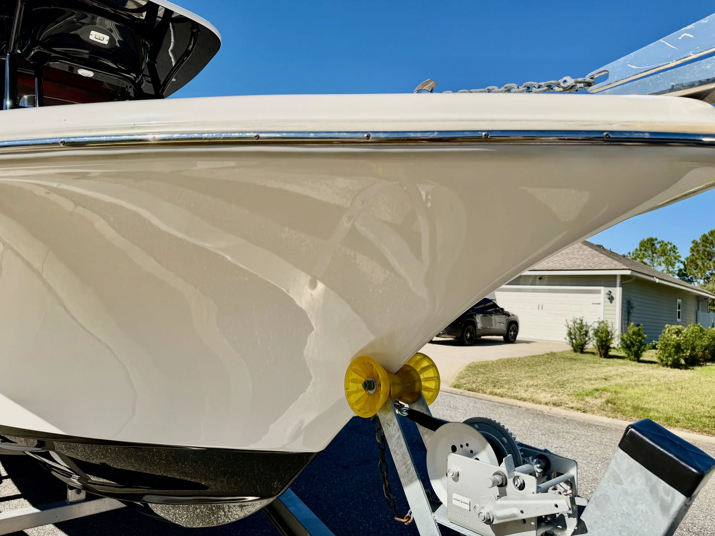 Close-up of the front of a white boat on a trailer, with the bow facing the camera, in a suburban driveway with a house, car, and trees in the background.