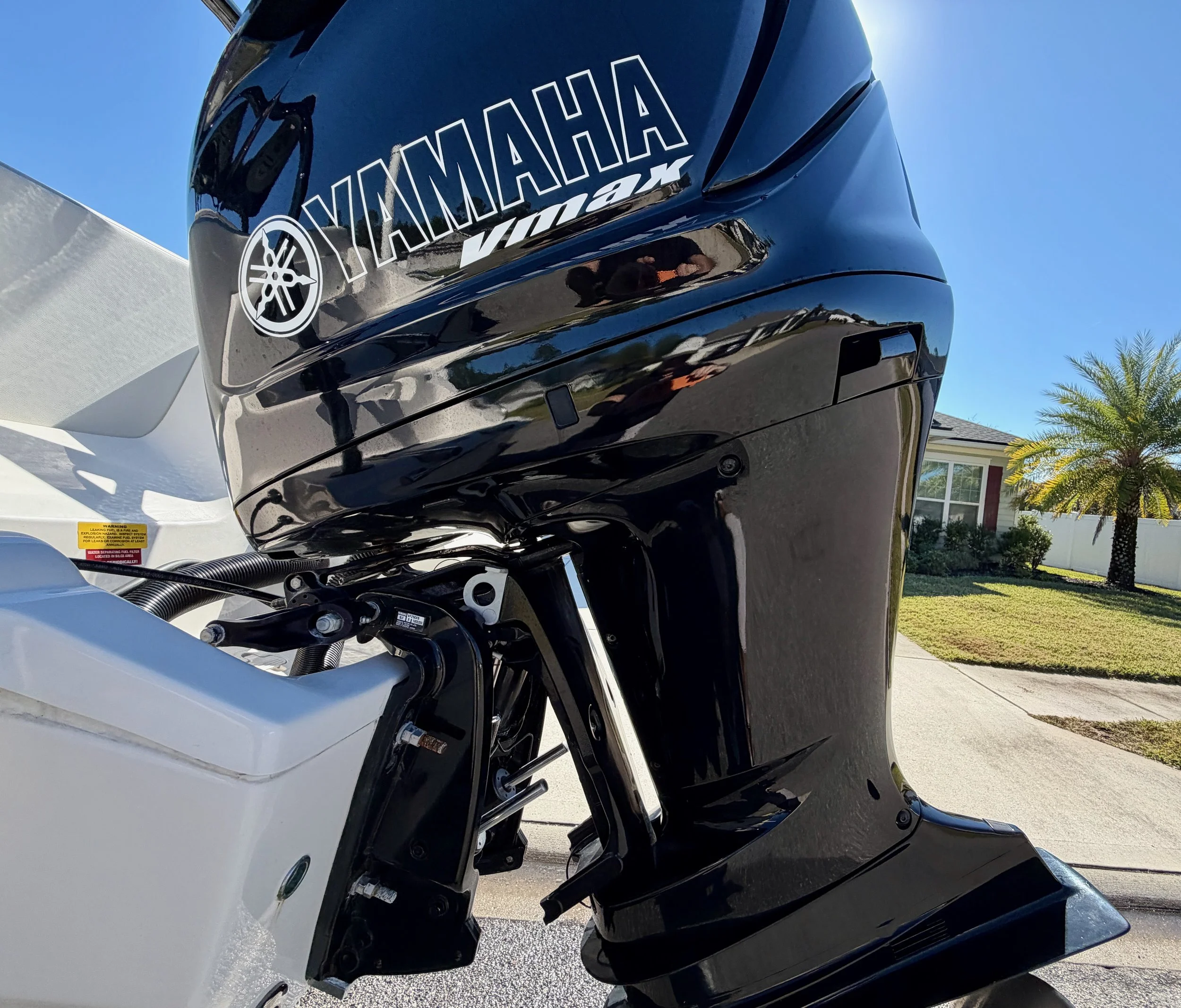 Close-up of a Yamaha outboard motor attached to a boat with a white hull, outdoors on a sunny day with a house, palm tree, and lawn in the background.