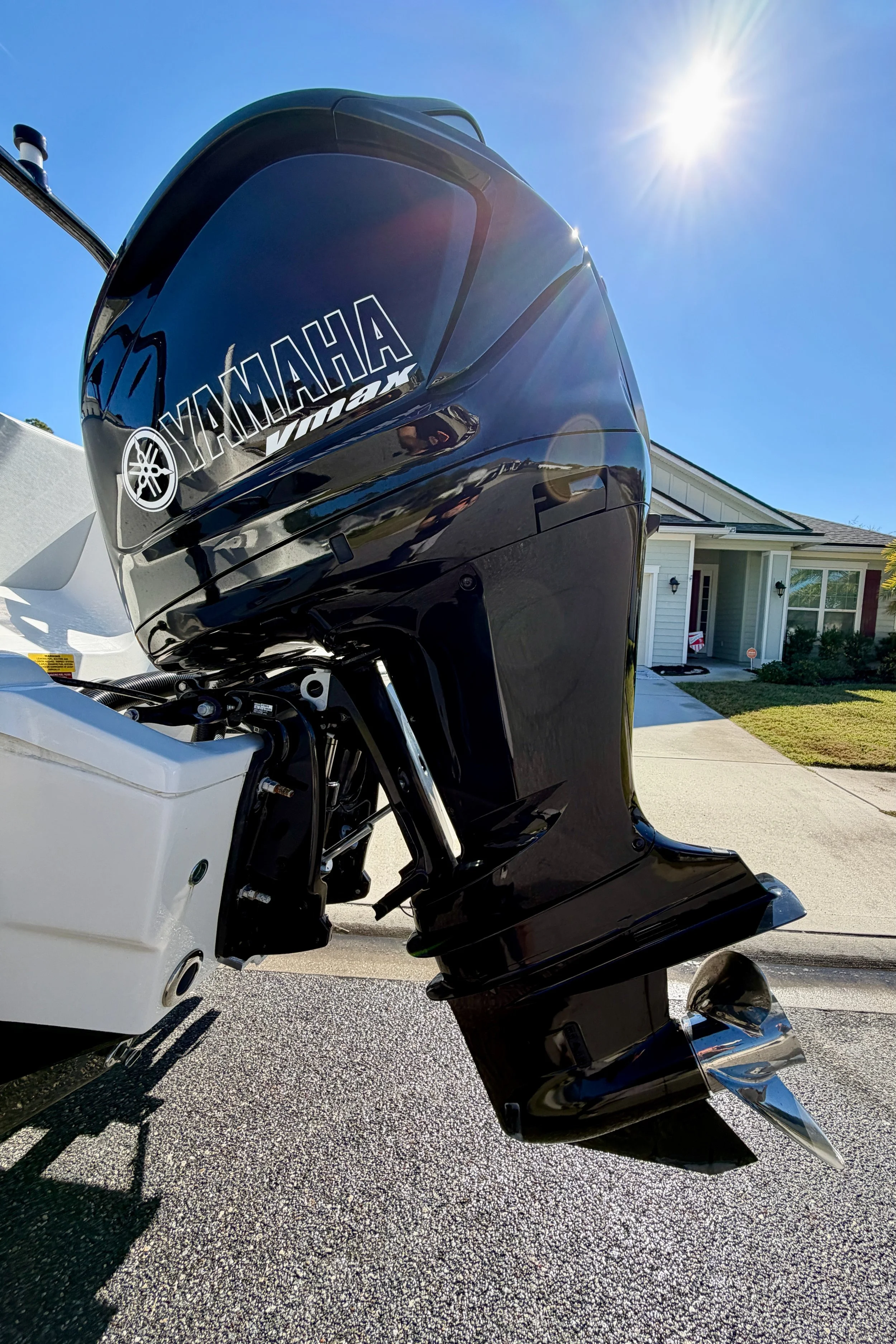 Close-up of a black Yamaha outboard motor attached to a boat, with a residential house and driveway in the background, under a clear blue sky with the sun shining.