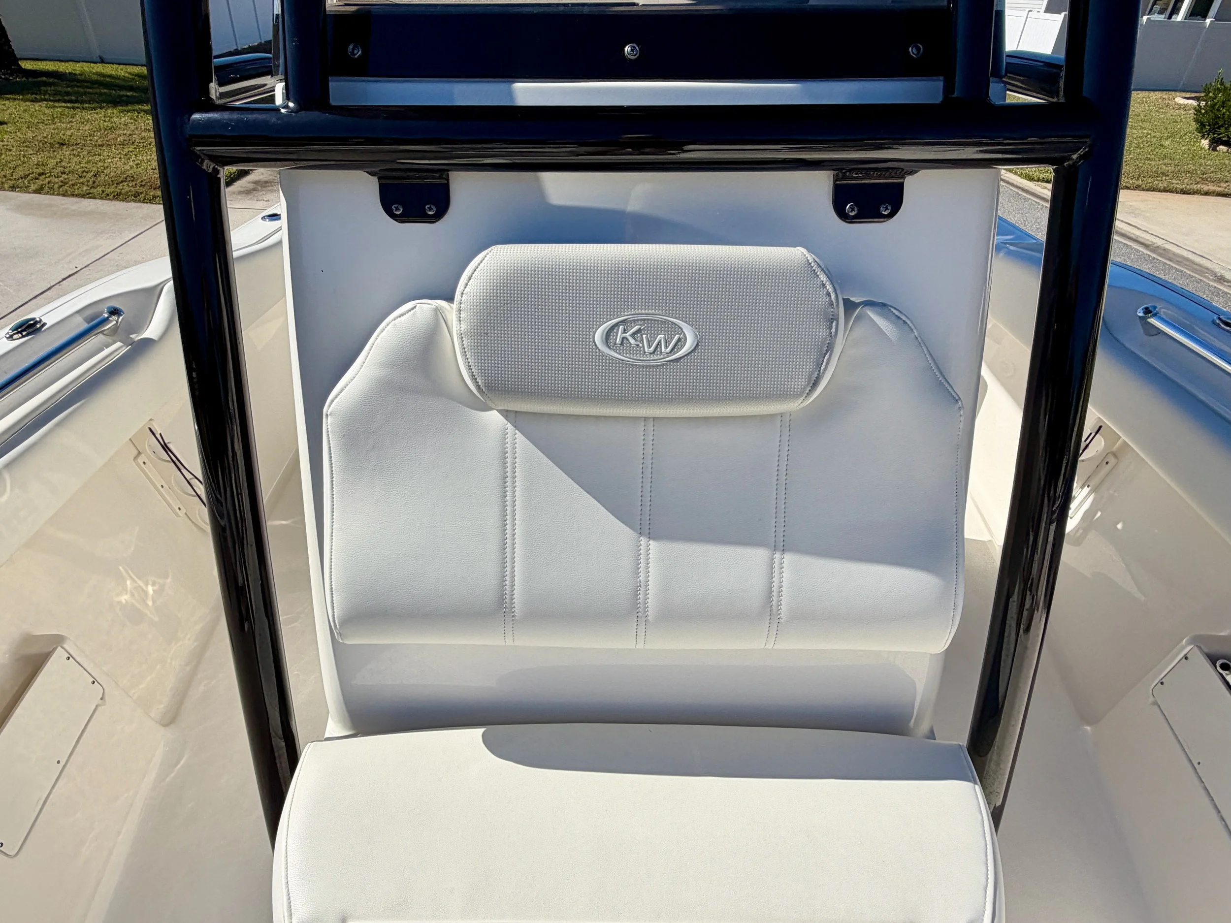 Close-up of a white boat seat with a headrest that has a KW logo, and a black metal frame, on a boat with a concrete walkway and grass in the background.