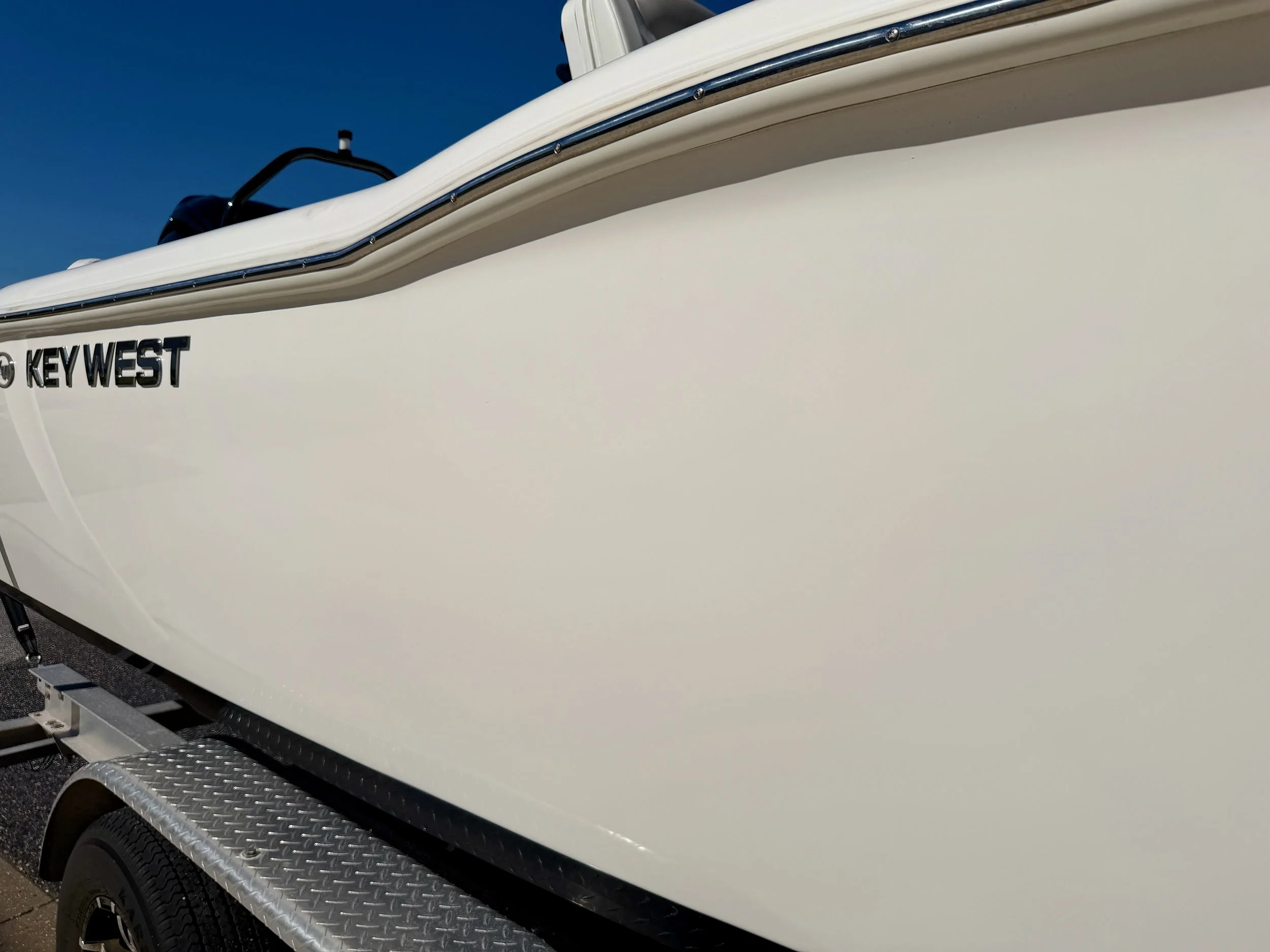Close-up of a white boat with the words 'KEY WEST' on its side, mounted on a trailer with a black tire and silver textured fender, against a clear blue sky.
