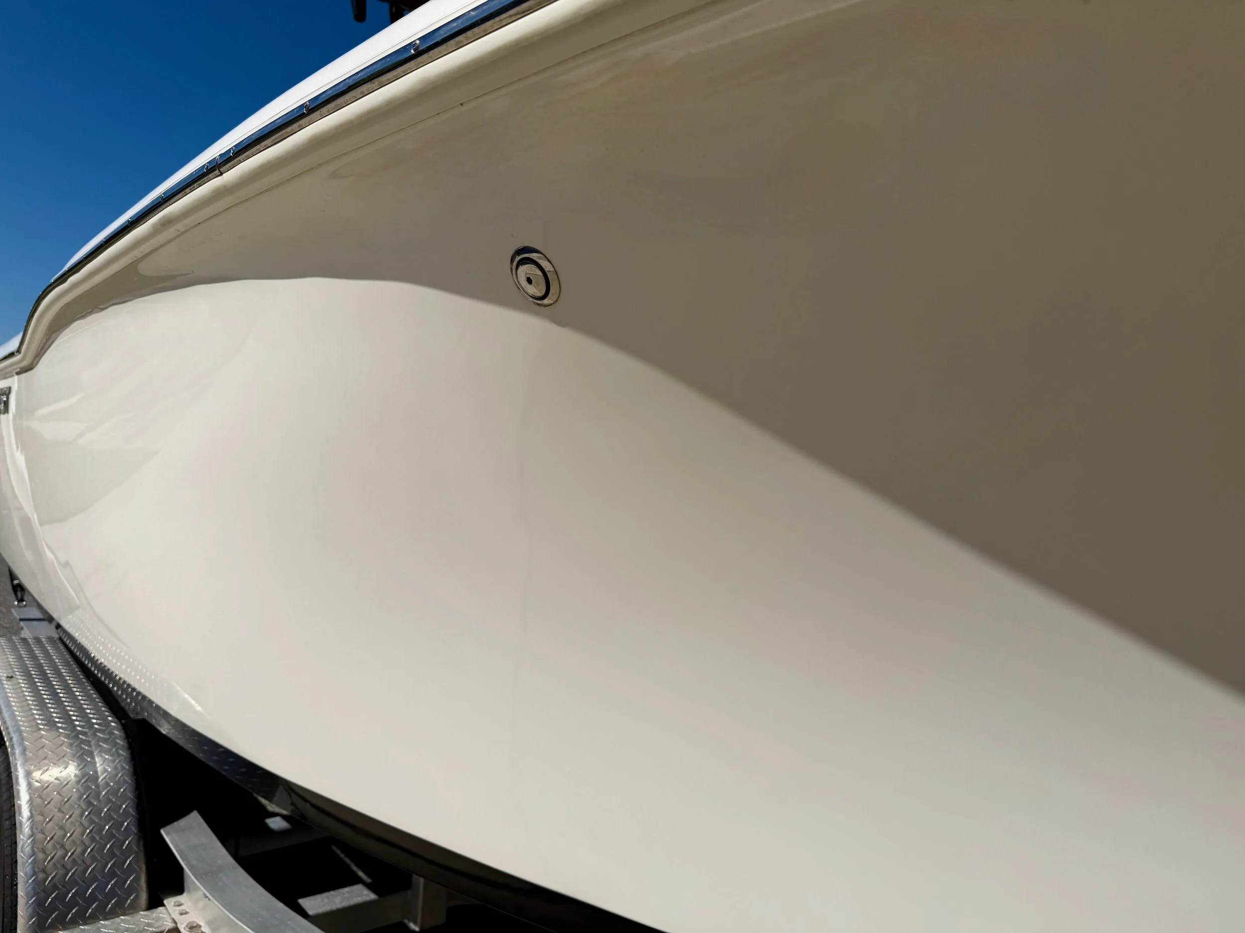 Close-up of a white boat's hull with a small inspection port, seen from below and slightly to the side, against a clear blue sky.