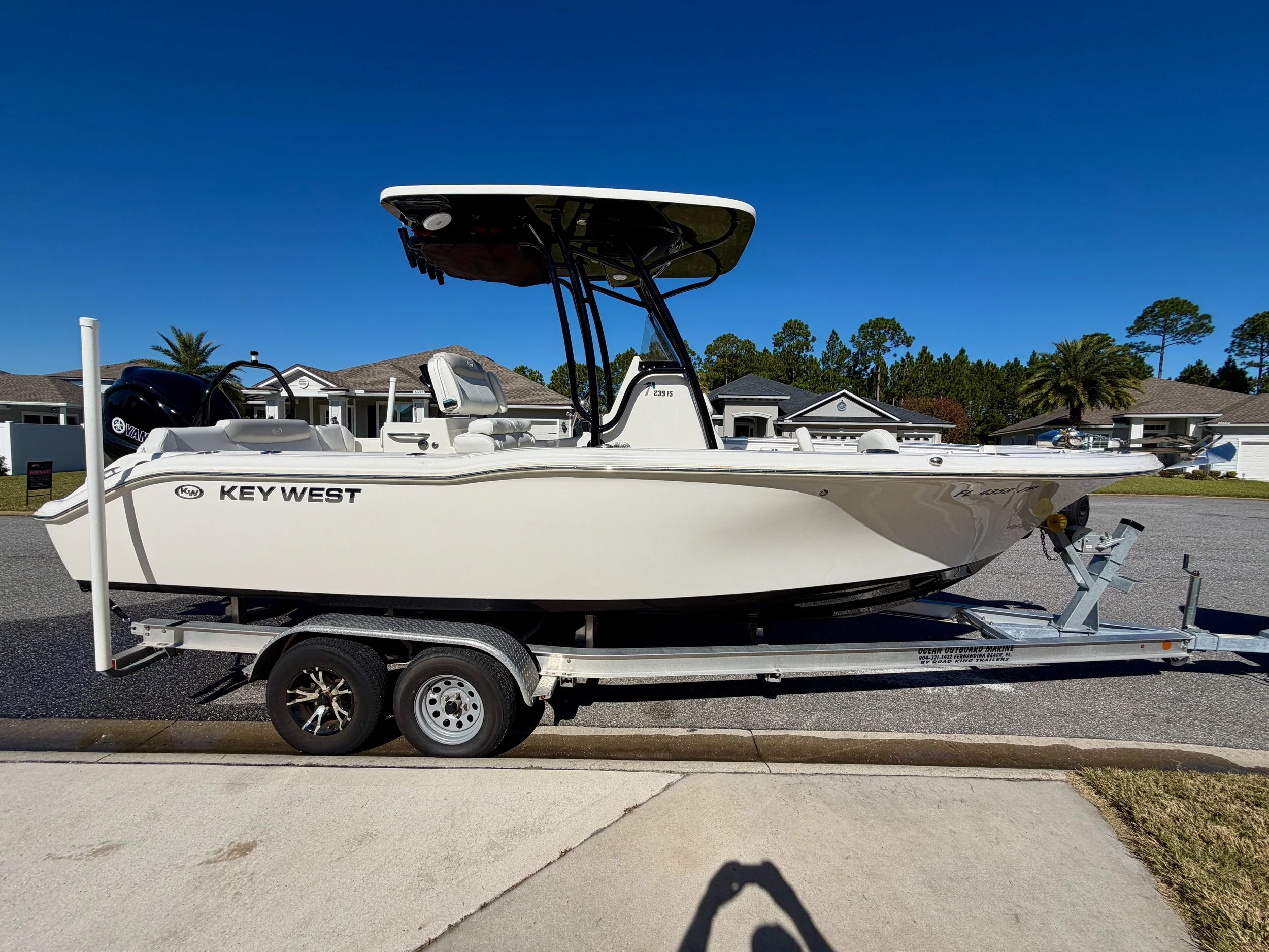 A white Key West boat on a trailer parked on the street in front of houses with palms and pine trees under a clear blue sky.