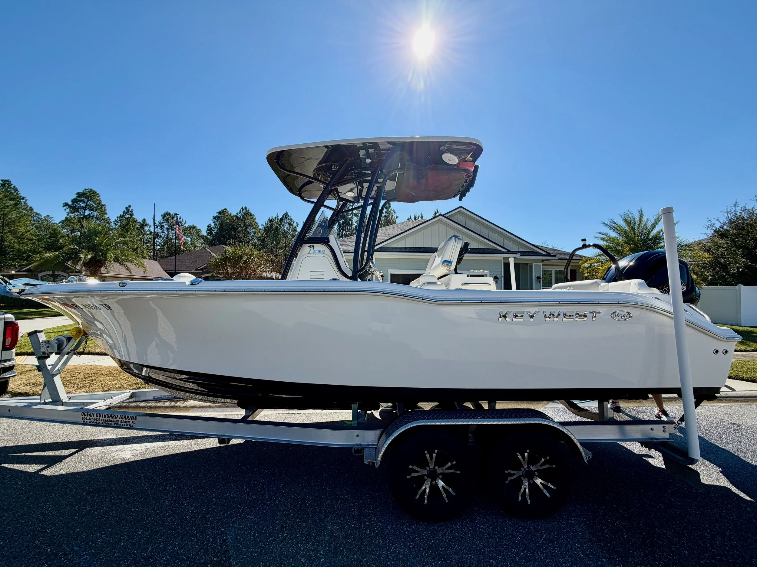 White Key West boat on a trailer, parked on a residential street with houses and trees in the background under a clear sunny sky.