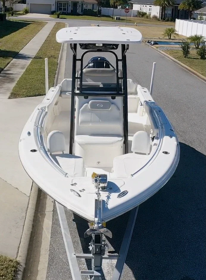 A white boat with a center console is parked on the street, attached to a trailer, with a suburban neighborhood and houses in the background.