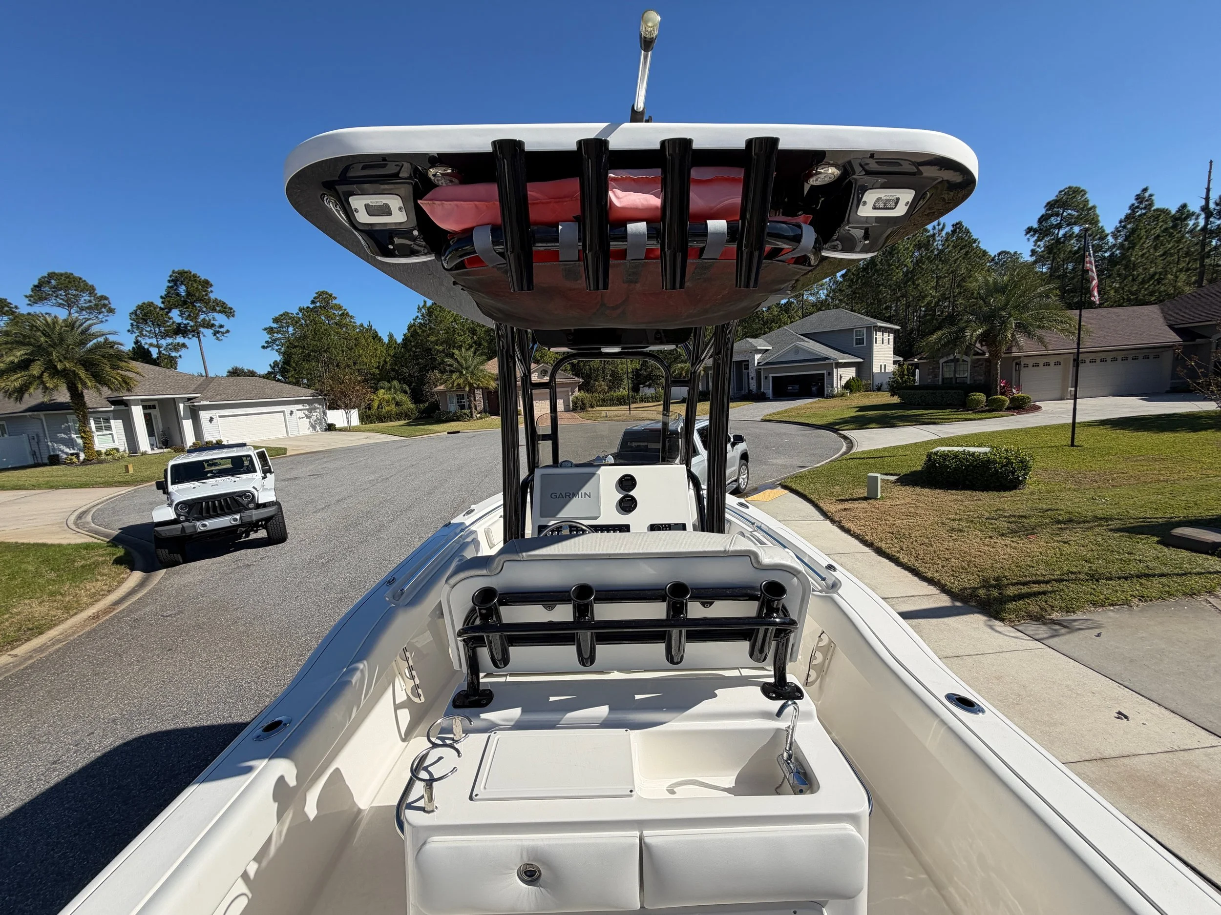View from the front of a fishing boat showing the T-top with mounted fishing rod holders, and a dashboard with electronic equipment, on a residential street with cars parked and houses in the background.