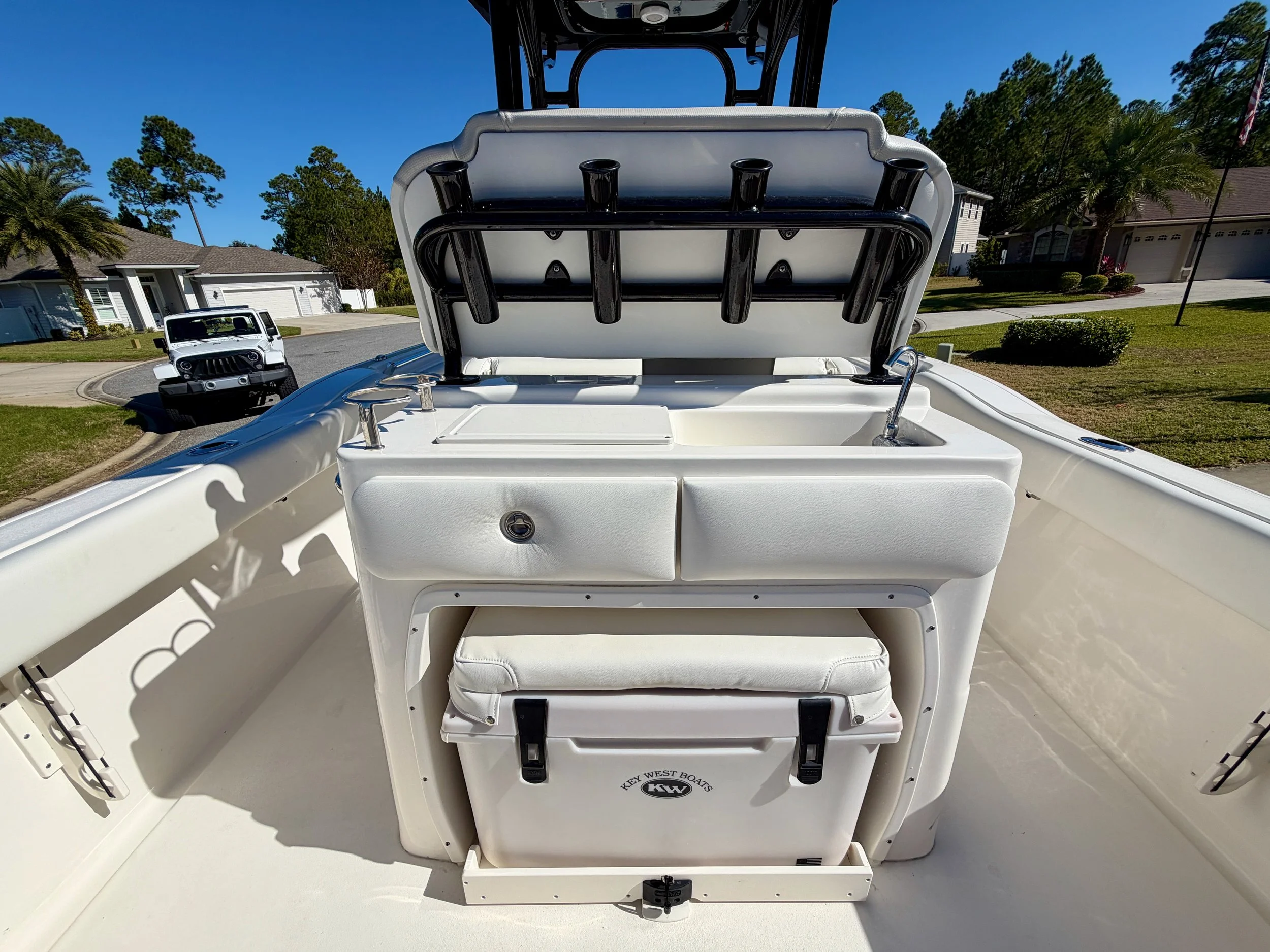 View of the bow of a white boat with a center console, cushioned seating, and a storage compartment, with a residential neighborhood and a Jeep in the background under a clear blue sky.