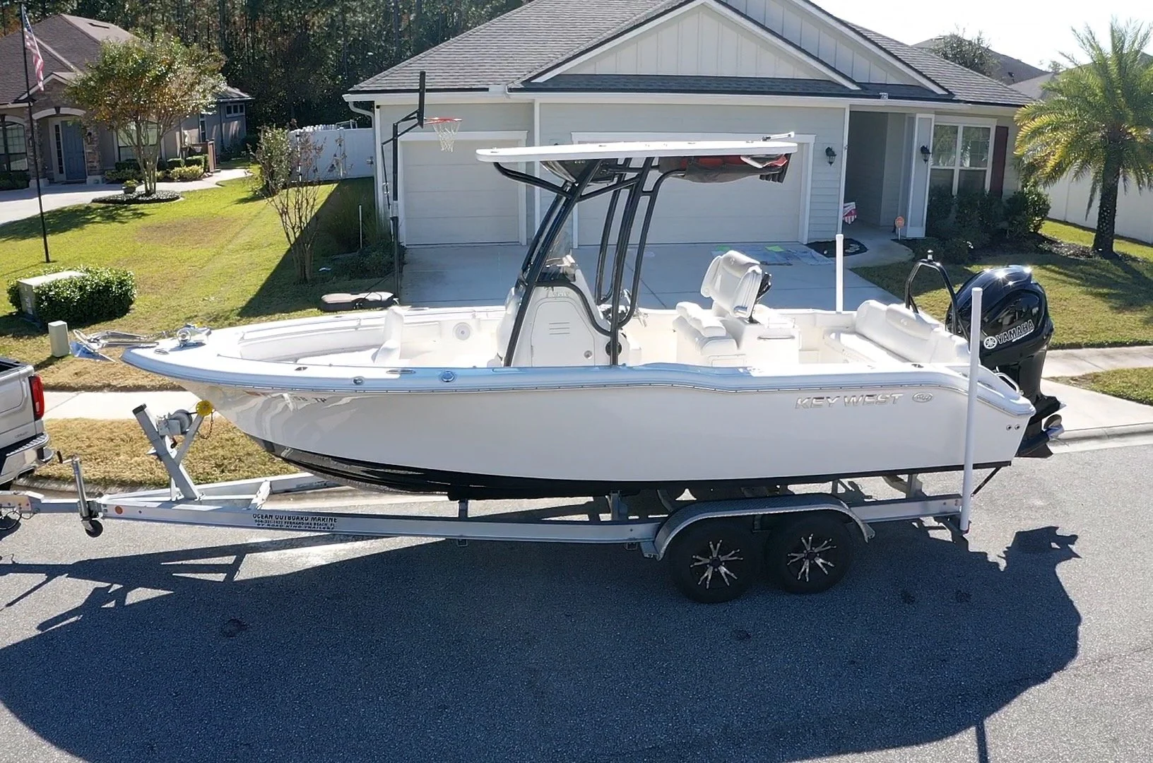 A white Key West center console boat on a trailer parked in a residential neighborhood street, with a garage and houses in the background, and an outboard Yamaha motor attached to the stern.