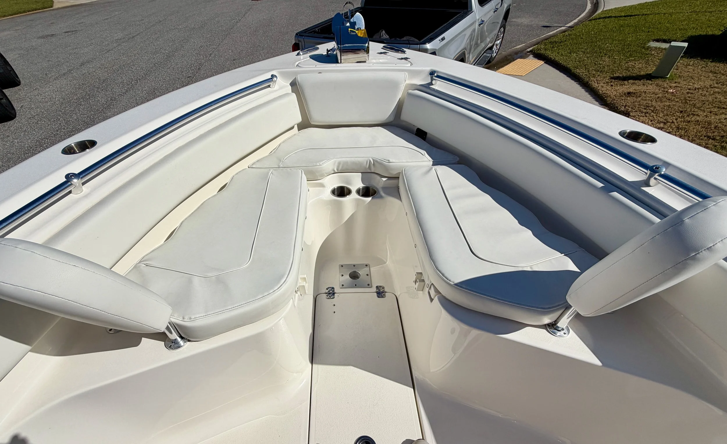The bow area of a white motorboat with cushioned seating and cup holders, parked near a street with a car and grass in the background.