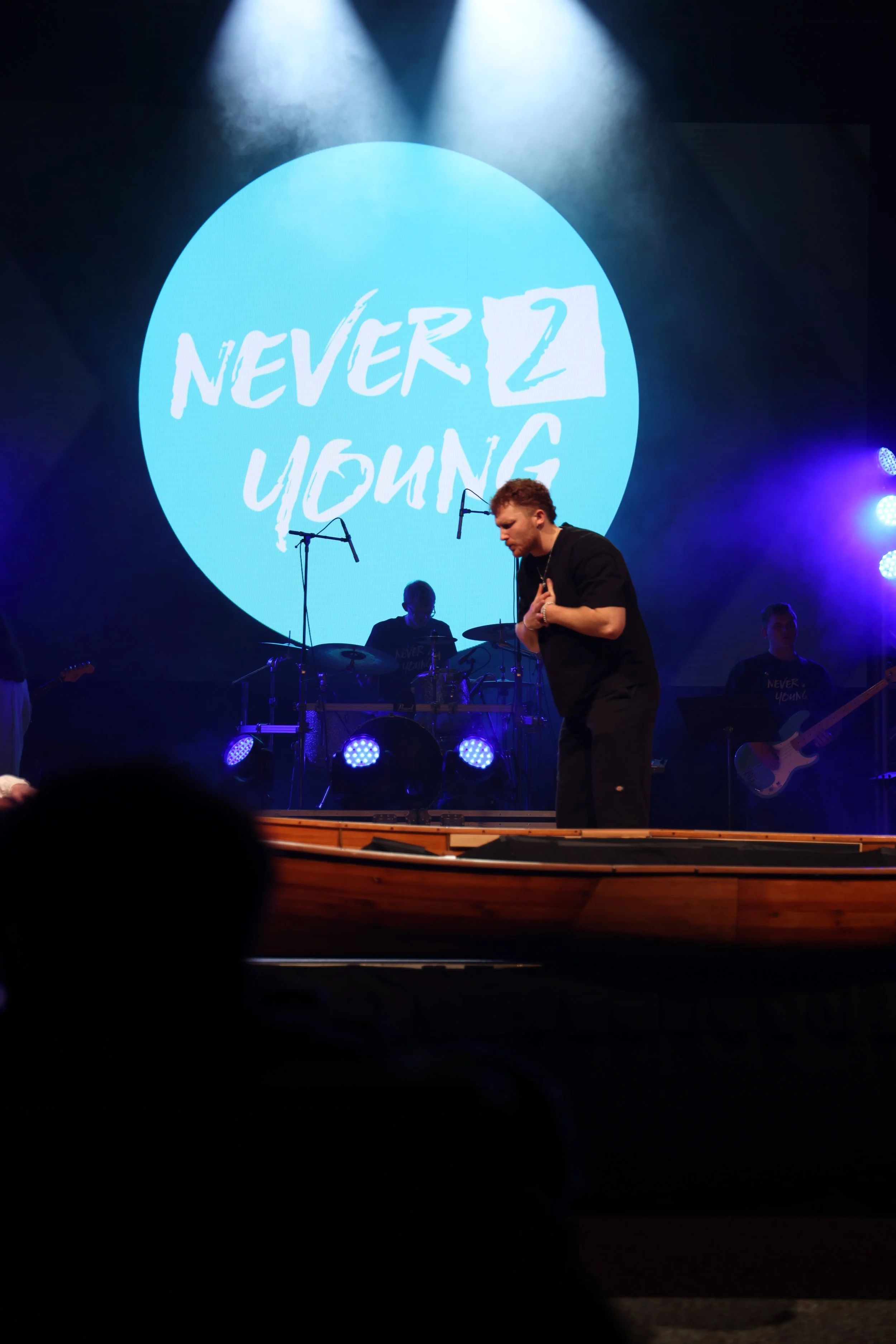 A live music performance on stage with a large screen behind displaying the words "NEVER 2 YOUNG" in a blue circle. A male singer is in front, holding his hand to his chest, with band members playing instruments in the background.
