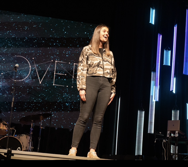 A woman standing on stage with a big screen behind her displaying the word 'LOVE' in a starry background. She is smiling and wearing a camouflage jacket and dark pants.