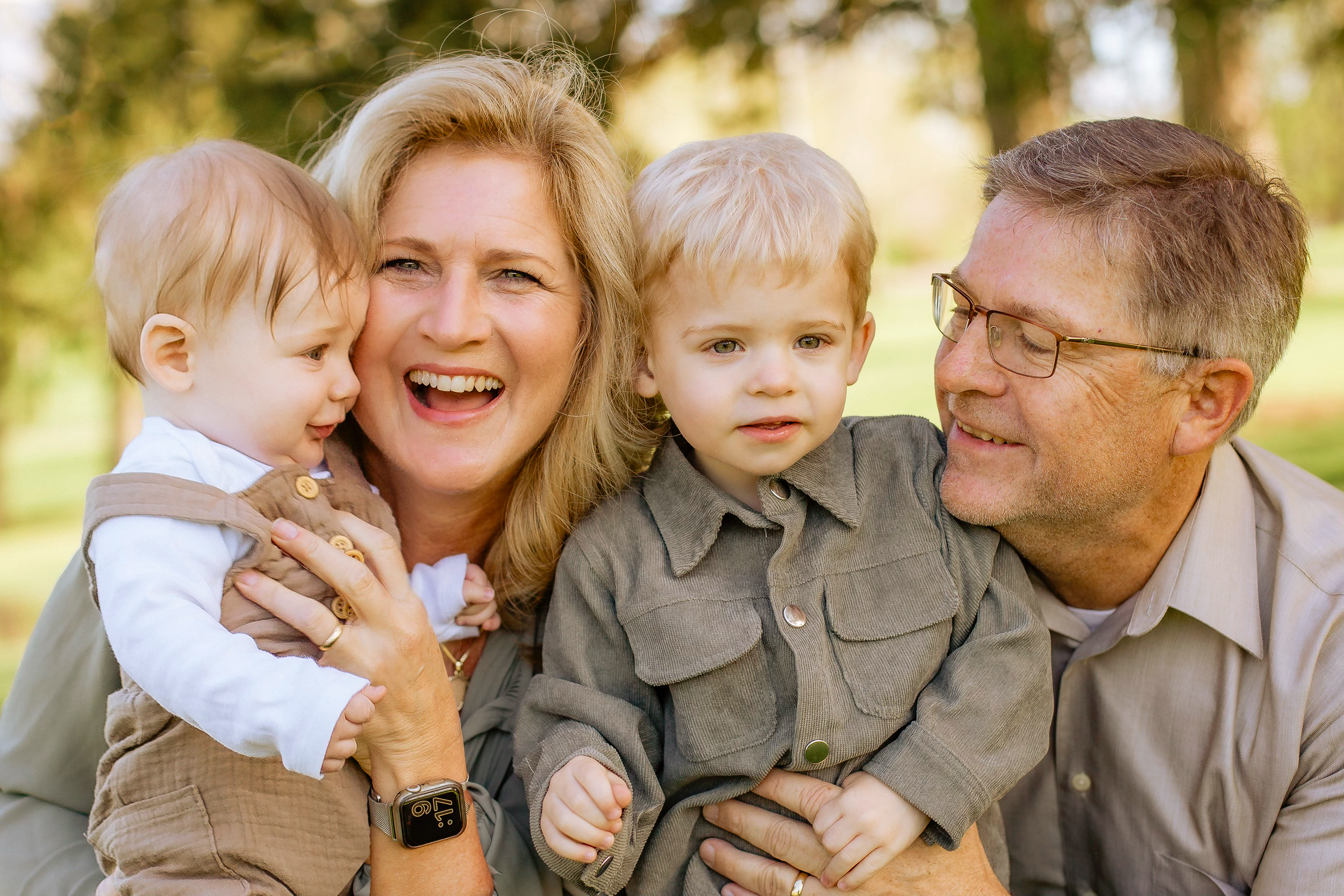 A smiling woman with blonde hair, holding a young boy, stands next to an older man with glasses, who is also holding a young boy. They are outdoors in a park with trees in the background.