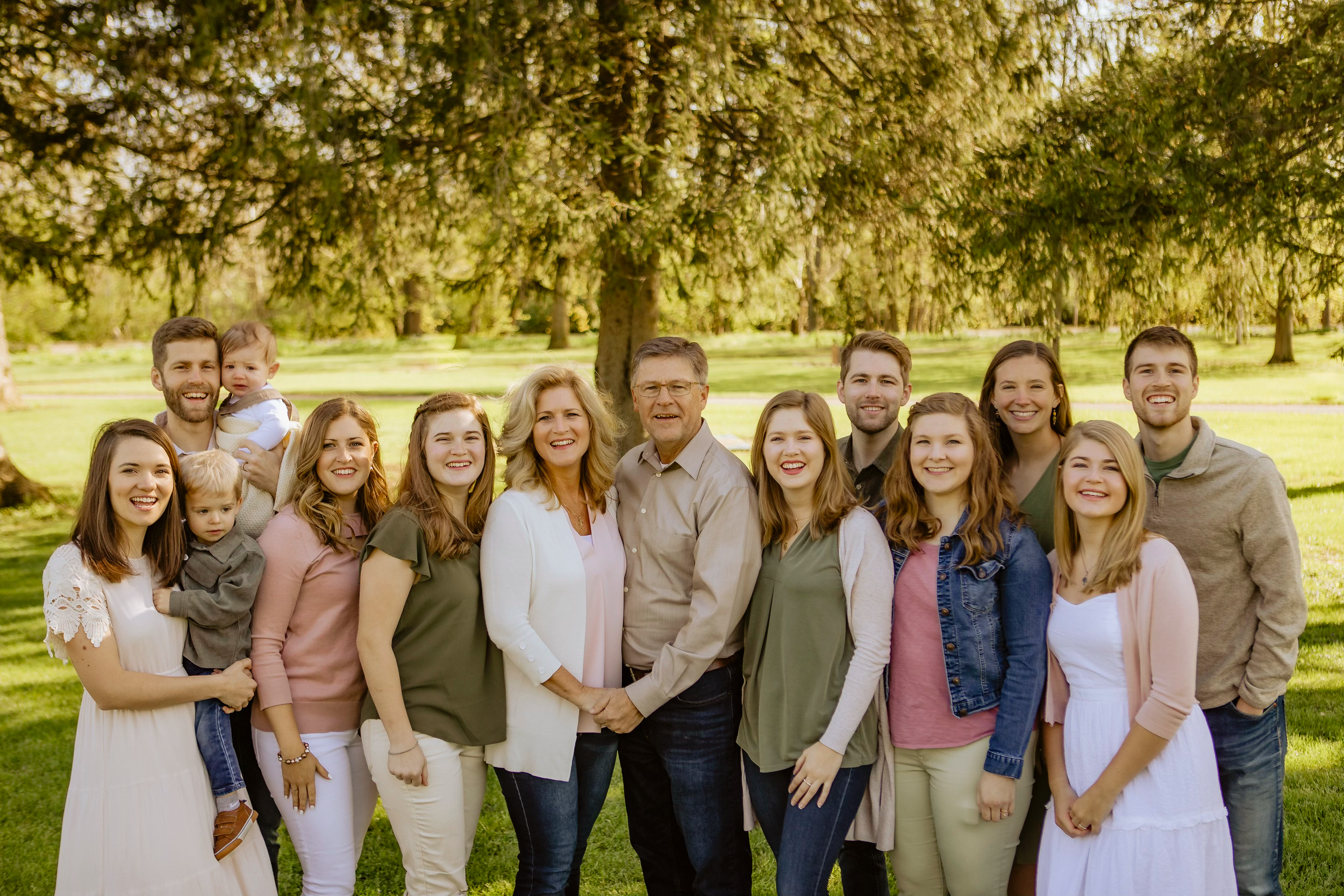 A large family group gathered outdoors in a park during daytime, smiling and posing for a photo with trees and grass in the background.