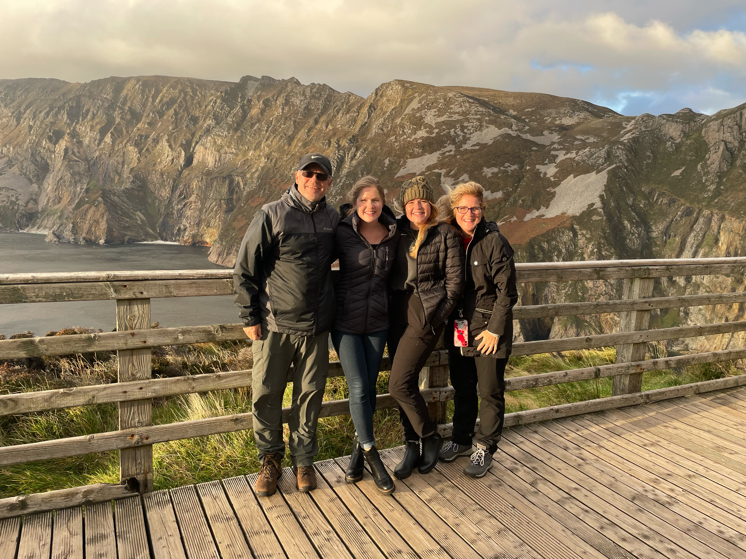 Four people standing on a wooden viewpoint with a scenic mountain landscape in the background, dressed in outdoor clothing.