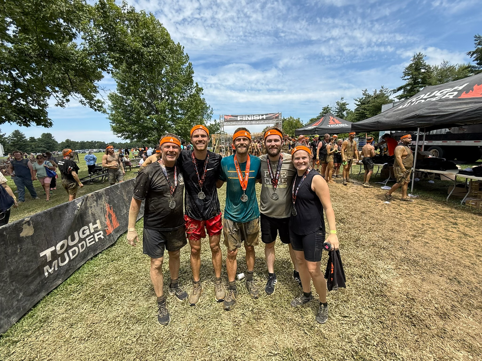 Group of five runners with medals celebrating after completing Tough Mudder obstacle course race, standing in front of the finish line with muddy clothes, orange headbands, and a muddy field background.
