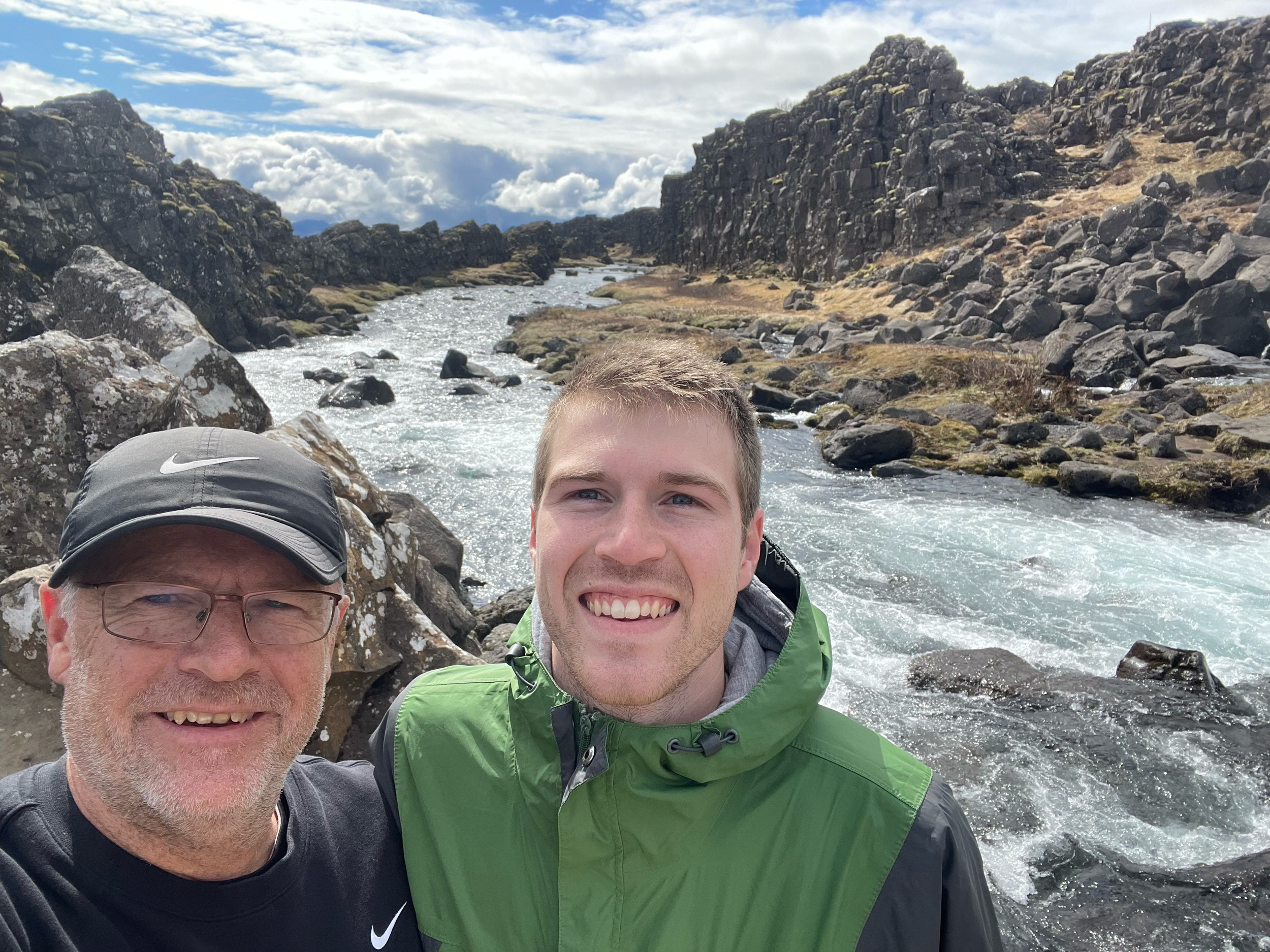 Two smiling men taking a selfie on rocky shoreline with ocean waves and cloudy sky in the background.