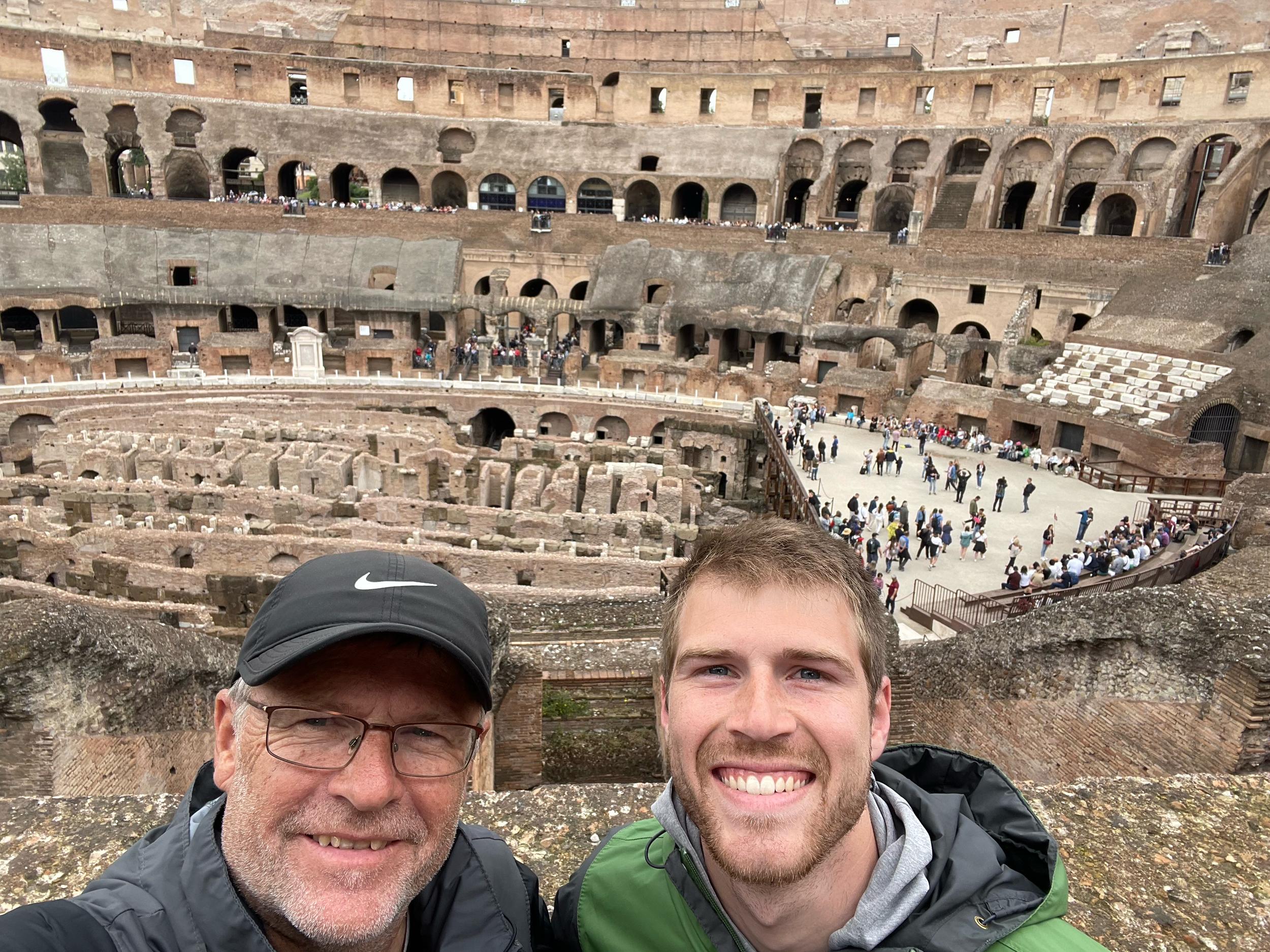Two smiling men taking a selfie inside the Colosseum in Rome, Italy, with the ancient amphitheater's interior and crowds of visitors visible in the background.