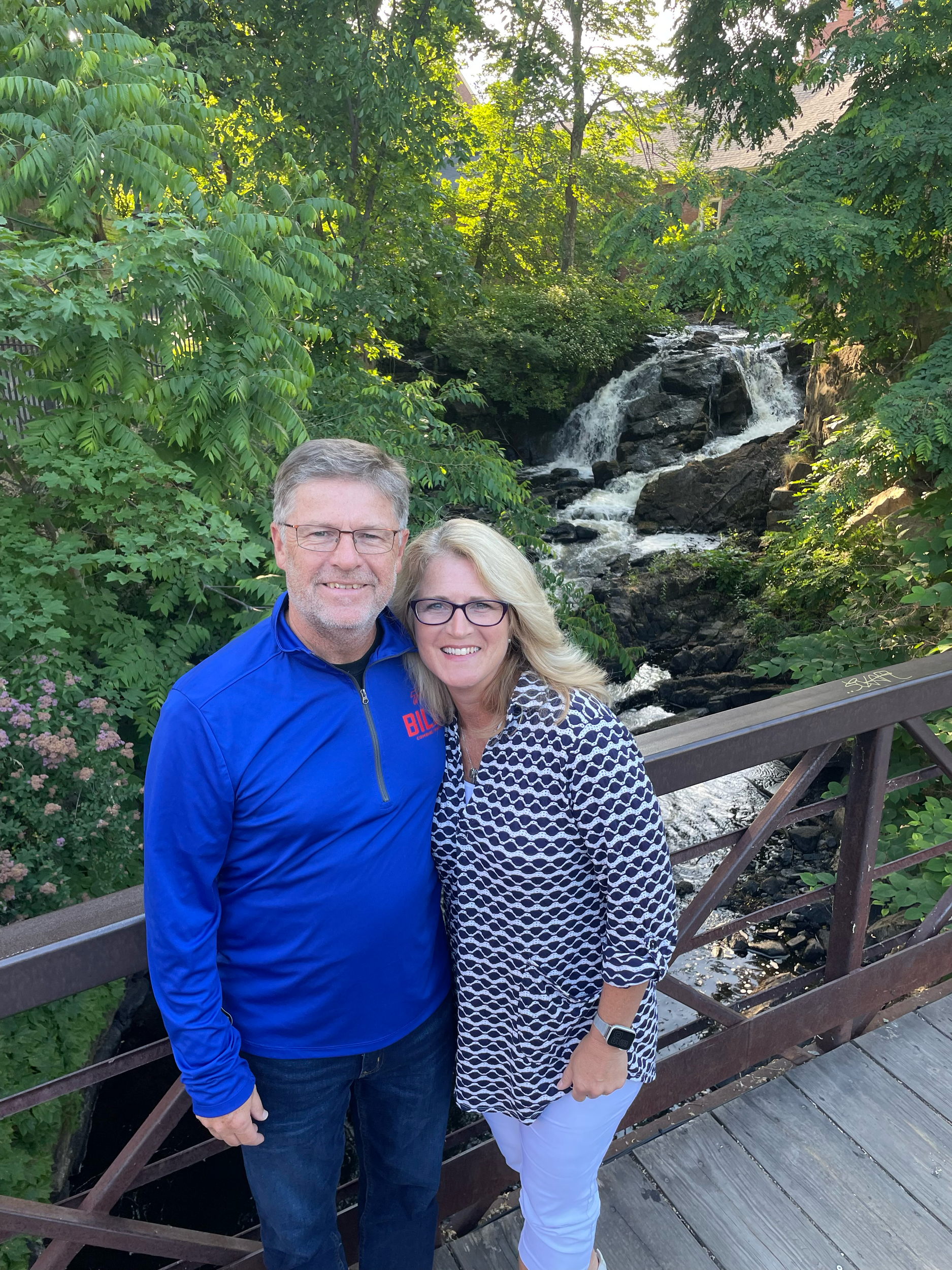 A smiling woman with blonde hair and glasses, wearing a patterned blouse and white pants, stands next to a smiling man with gray hair and glasses, wearing a blue jacket, on a wooden bridge with a small waterfall and lush green foliage in the backgrou