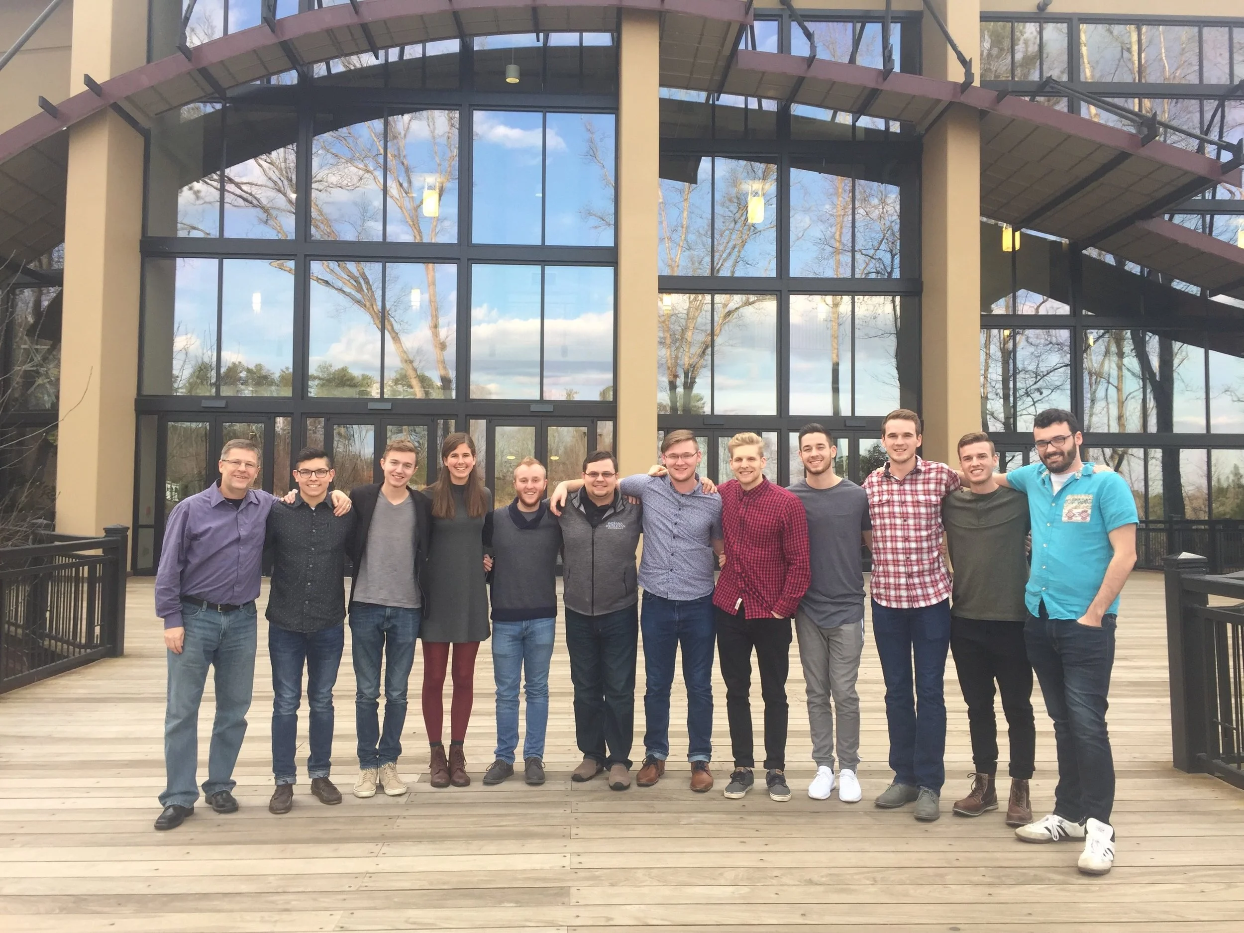 A group of 13 young adults standing arm-in-arm on a wooden deck in front of a large glass building with trees and sky reflected in the windows.