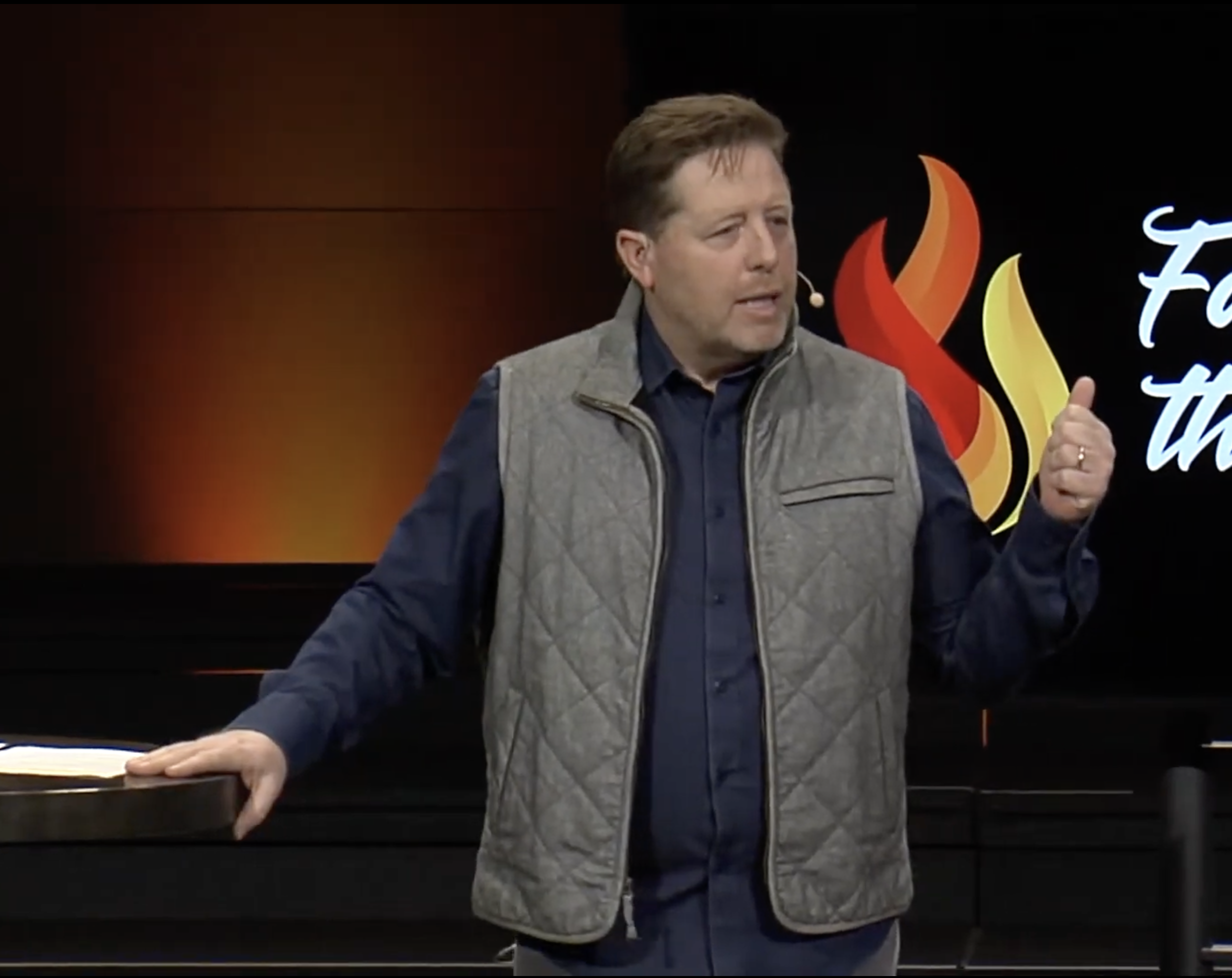 A man in a gray vest and dark blue shirt speaking on stage with a microphone headset, gesturing with his right hand, standing in front of a black background with a logo that has flames and text.