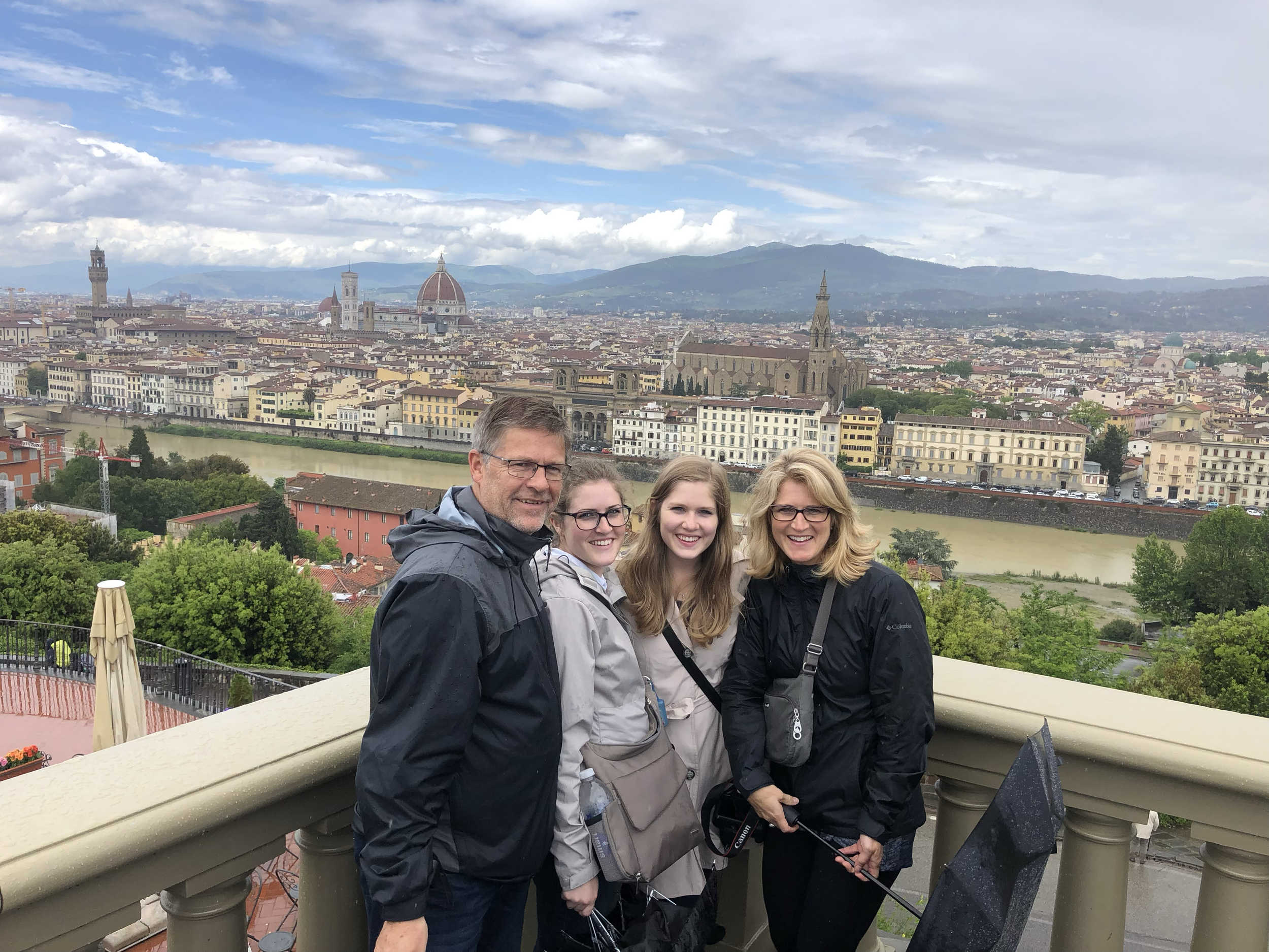 Four people smiling on a balcony with a cityscape of Florence, Italy, including the Florence Cathedral, River Arno, and rolling hills in the background.