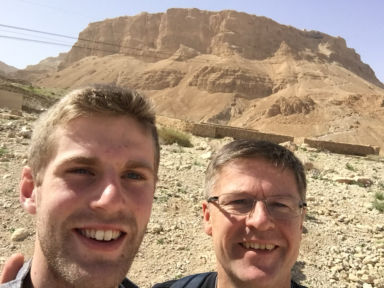 Two men smiling for a selfie in a rocky desert landscape with a large, layered mountain in the background.