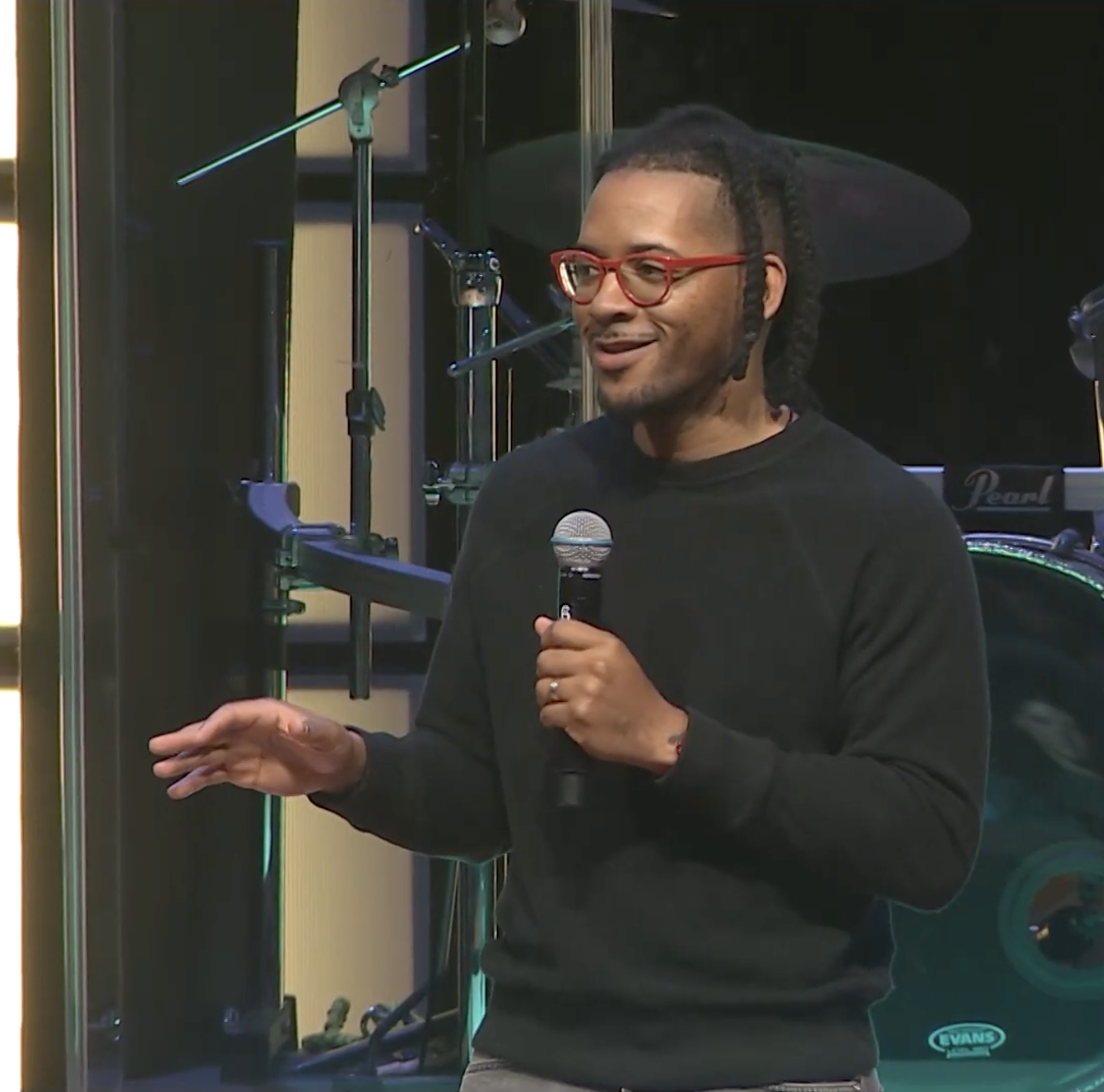 A man with glasses speaking into a microphone in front of a musical drum set.