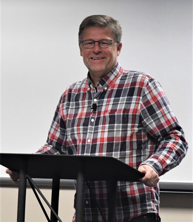 A middle-aged man with glasses and short gray hair stands at a podium, smiling, wearing a plaid shirt with red, black, and white colors, in a room with a whiteboard or projection screen behind him.