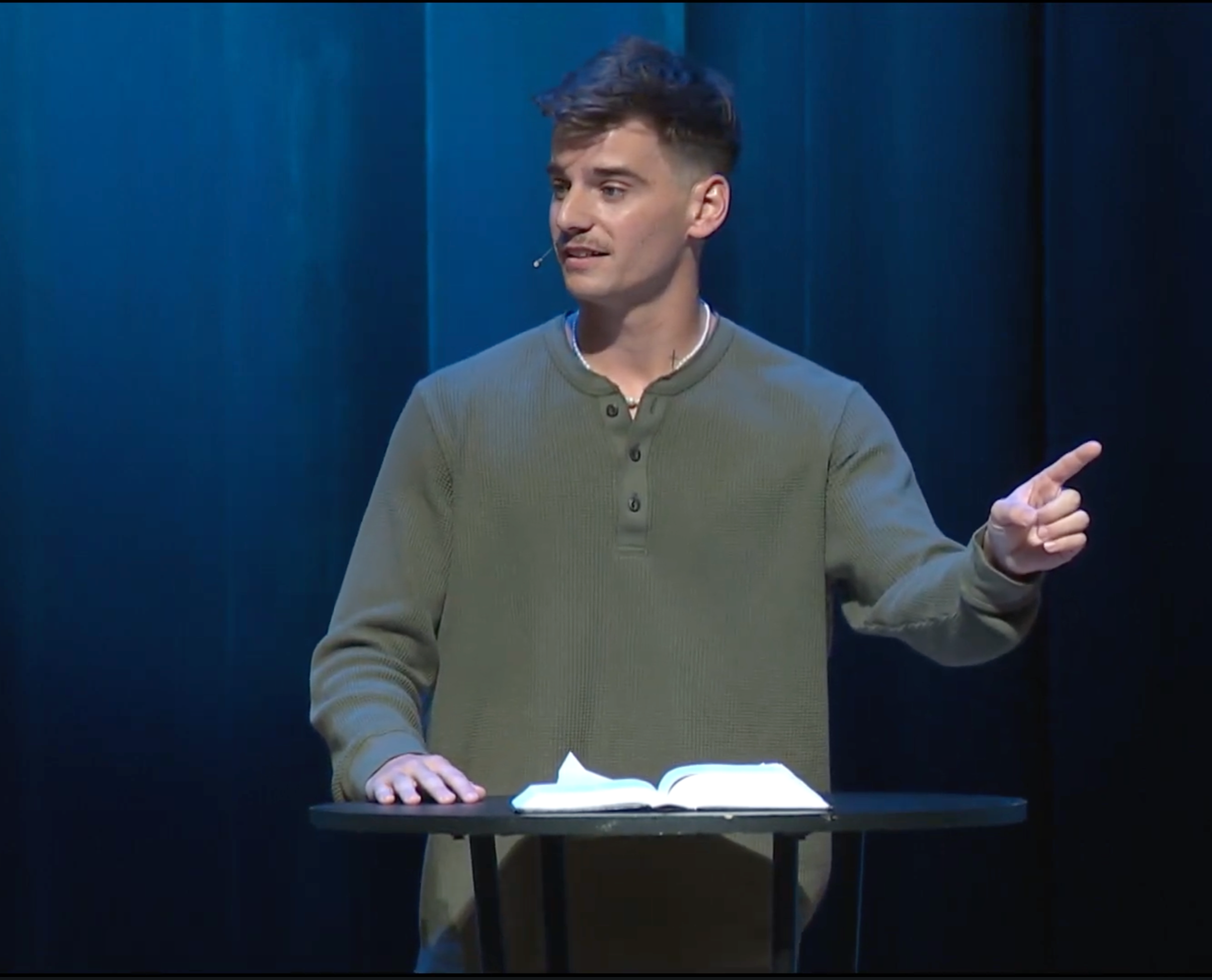 A young man with brown hair wearing a green long-sleeve shirt standing behind a small black table with an open book on it, gesturing with his right hand, and speaking into a microphone.