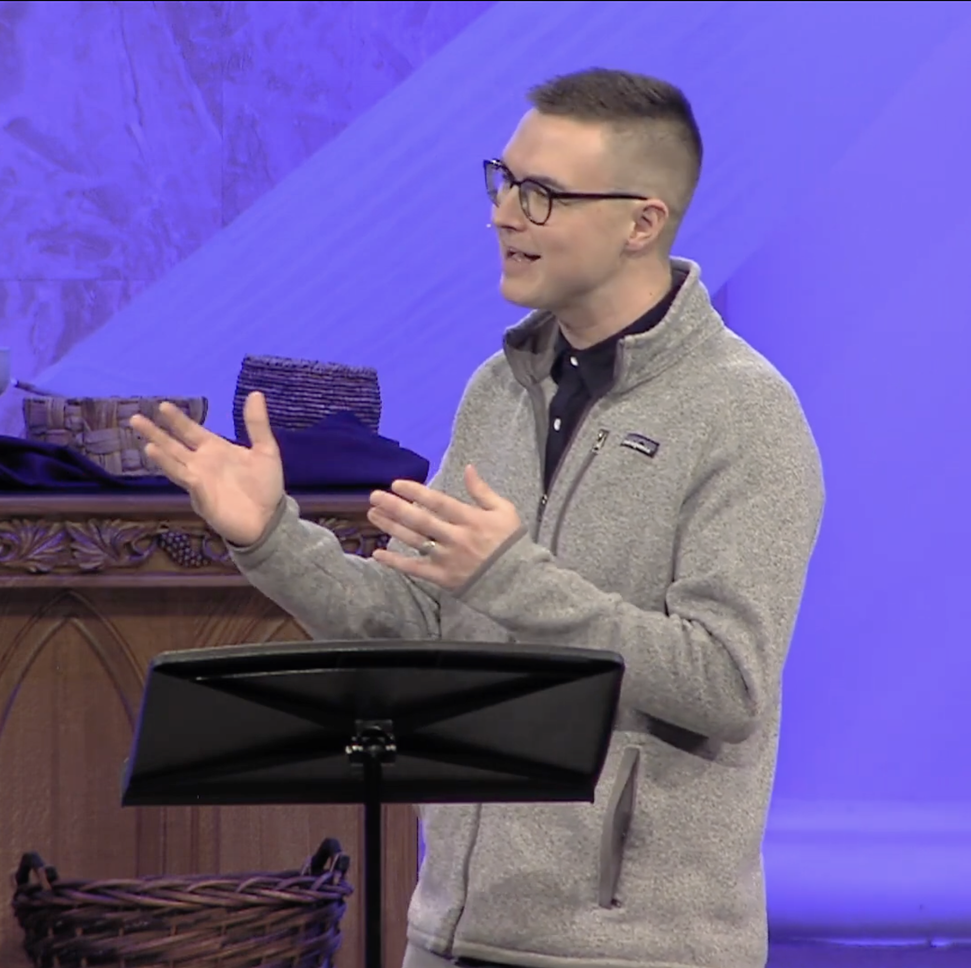 A man with glasses and short hair, wearing a gray jacket, is talking and gesturing with his hands. There is a music stand in front of him and a woven basket and dark folded textiles on a wooden surface behind him. The background features a wall with purple and blue lighting.