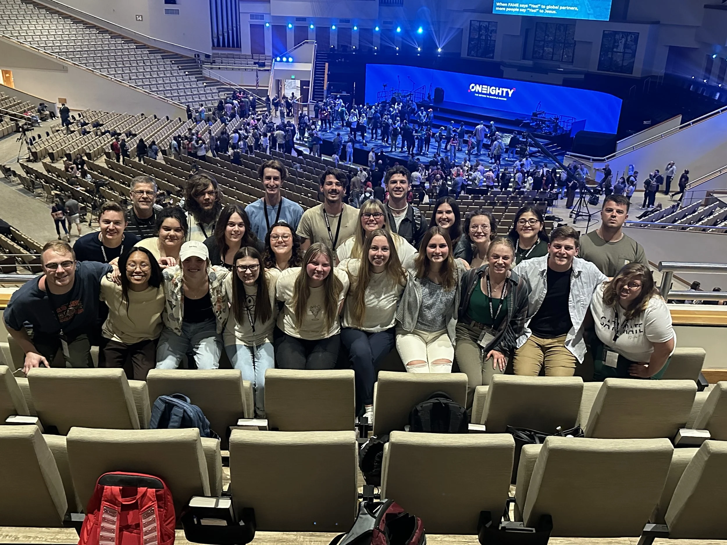 A group of people posing together in an auditorium with many empty seats, a stage, and a large screen displaying the words 'ONEighty.' The background shows a crowd on the stage preparing for an event.