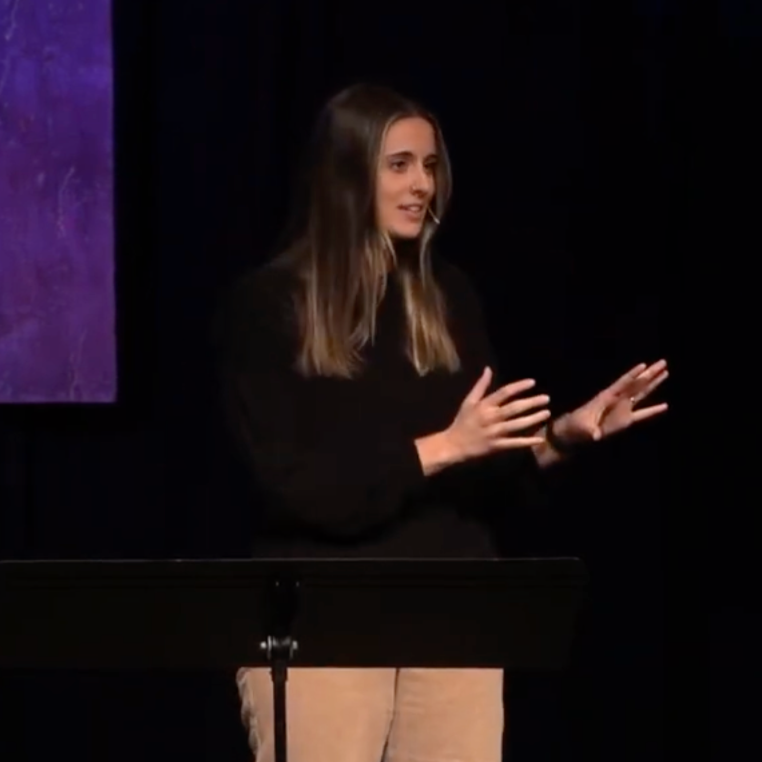 Young woman speaking or presenting on a stage, gesturing with her hands, standing behind a black music stand, with a dark background.