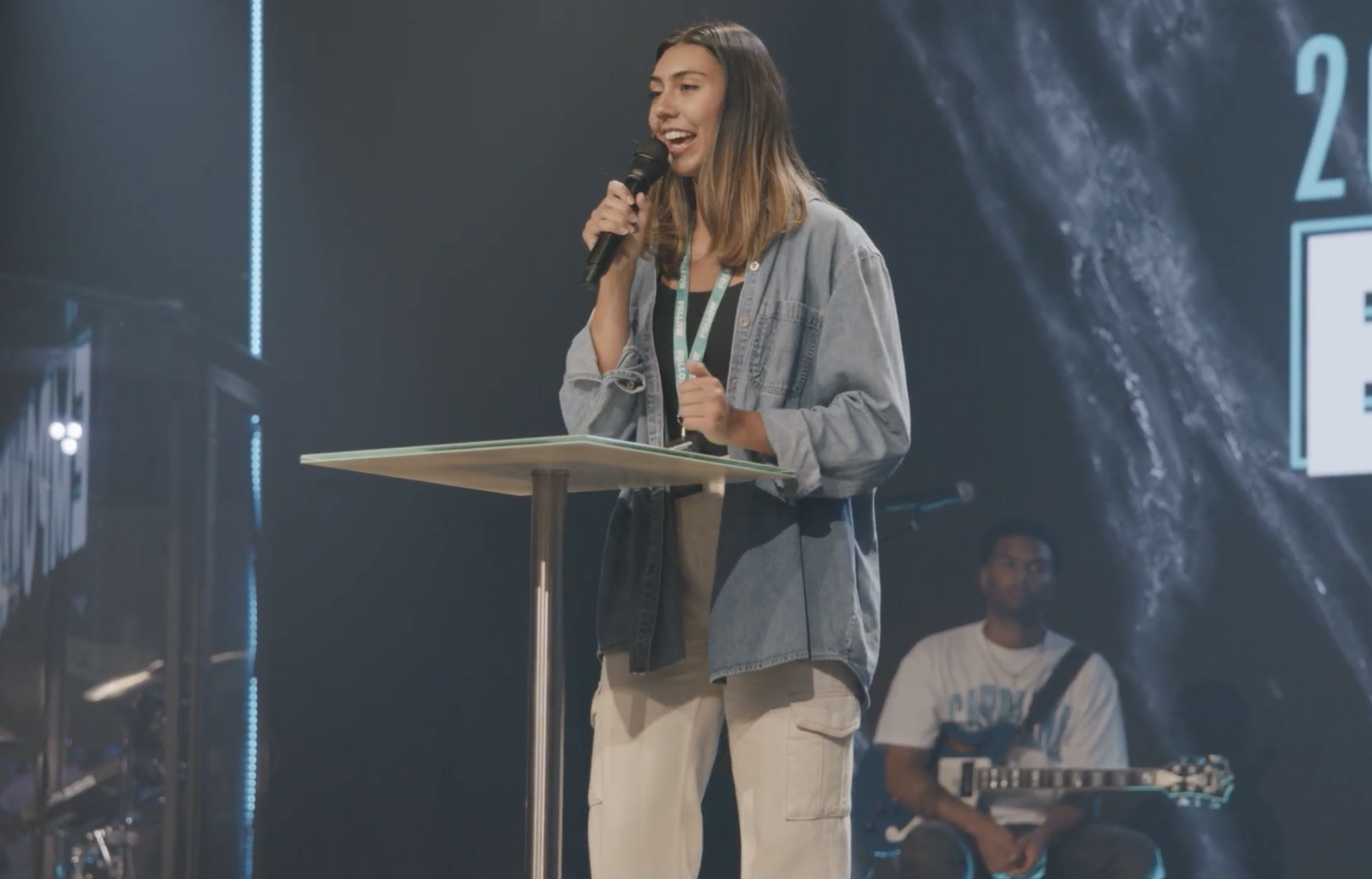 A woman with long brown hair, wearing a gray denim jacket and beige cargo pants, speaking into a microphone at a podium during a presentation or event. Behind her, a man with a beard, wearing a white T-shirt with a graphic and holding an electric guitar, is seated.