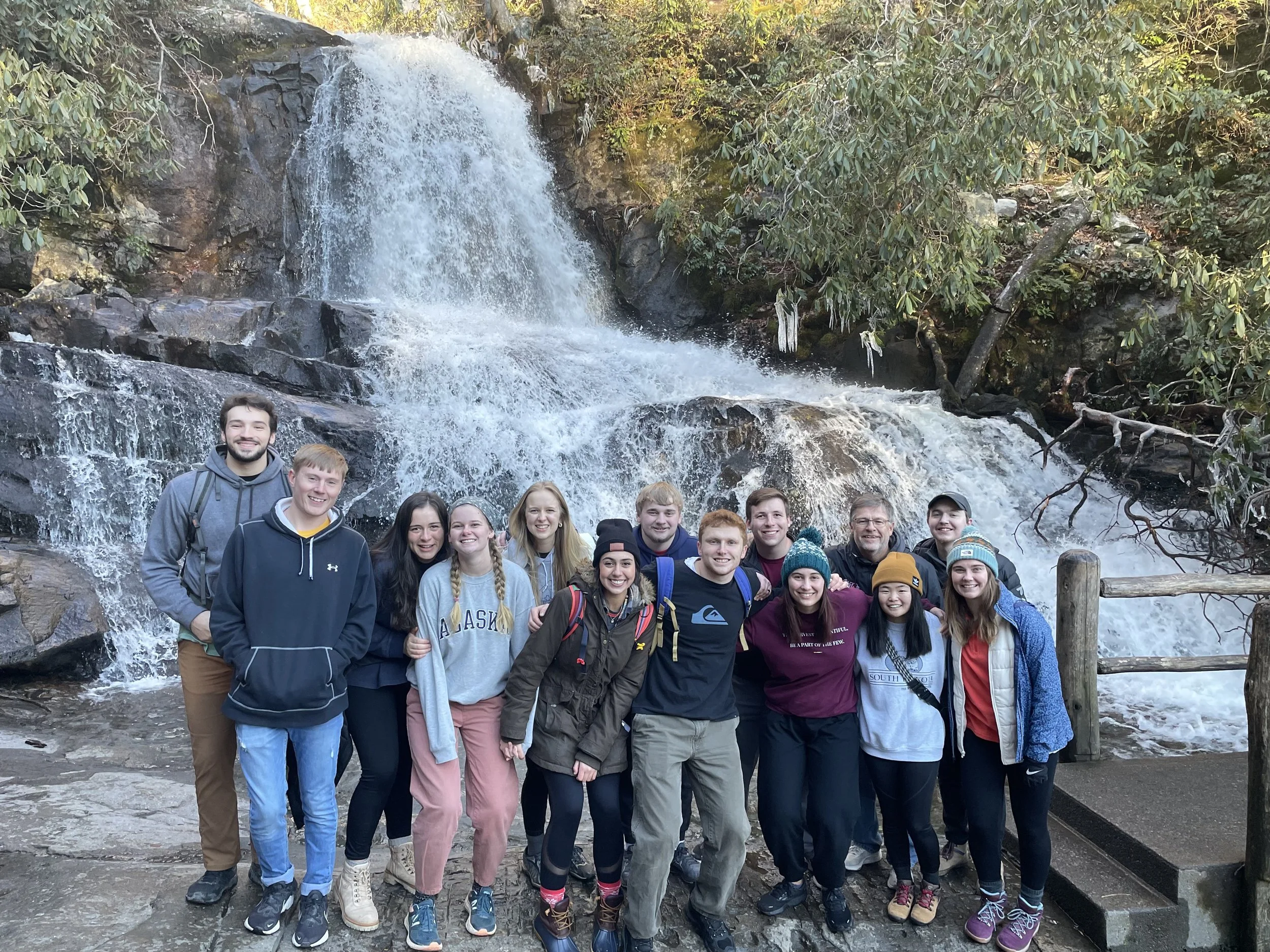 Group of 14 people standing in front of a waterfall during a hike. They are smiling and dressed in casual outdoor clothing, some wearing hats and backpacks.
