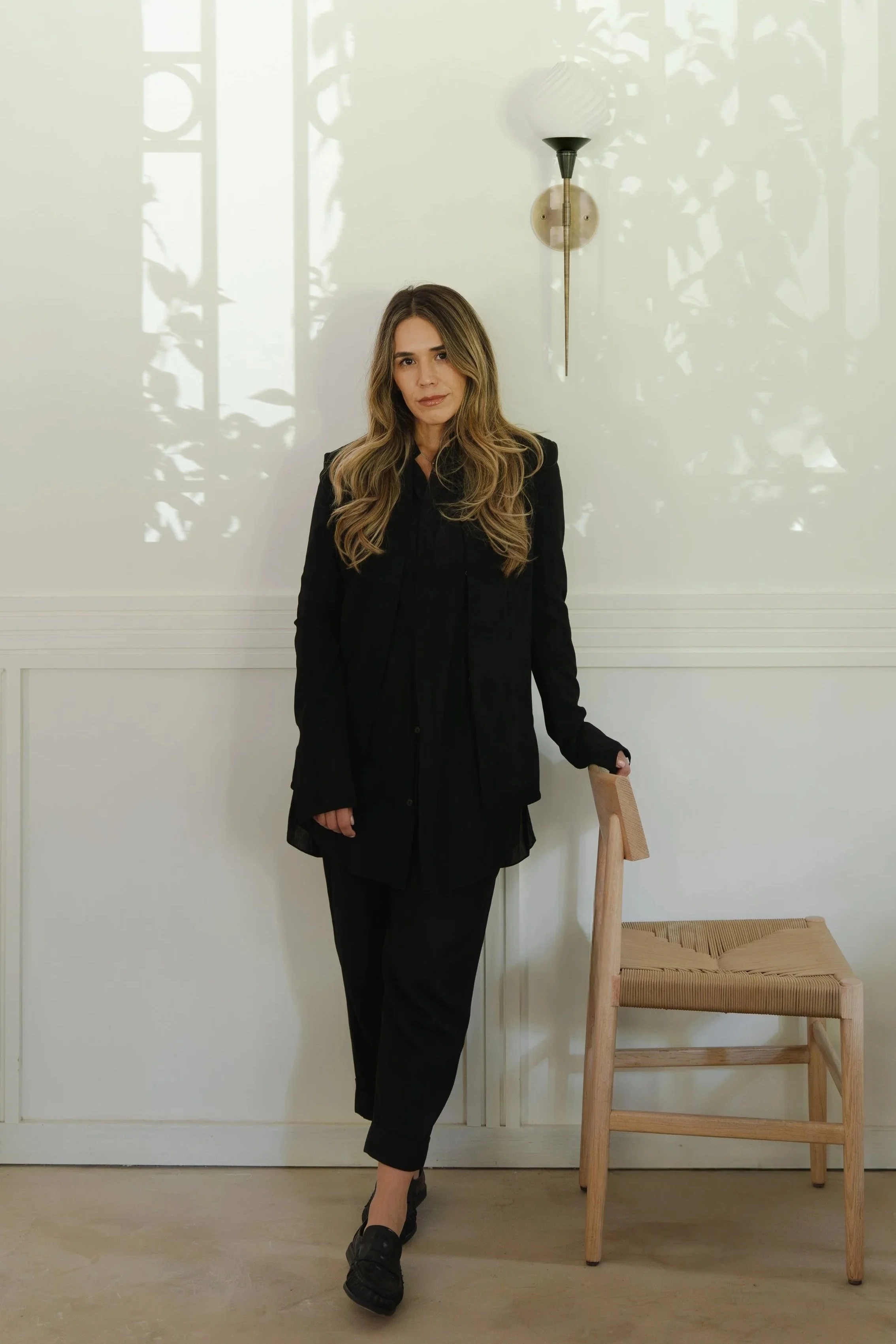 A woman with long, wavy hair standing next to a wooden chair with a woven seat. She is dressed in black clothing, including a blazer and pants, and is wearing black shoes. Behind her is a white wall with a sconce light fixture and shadows of plants cast on the wall.