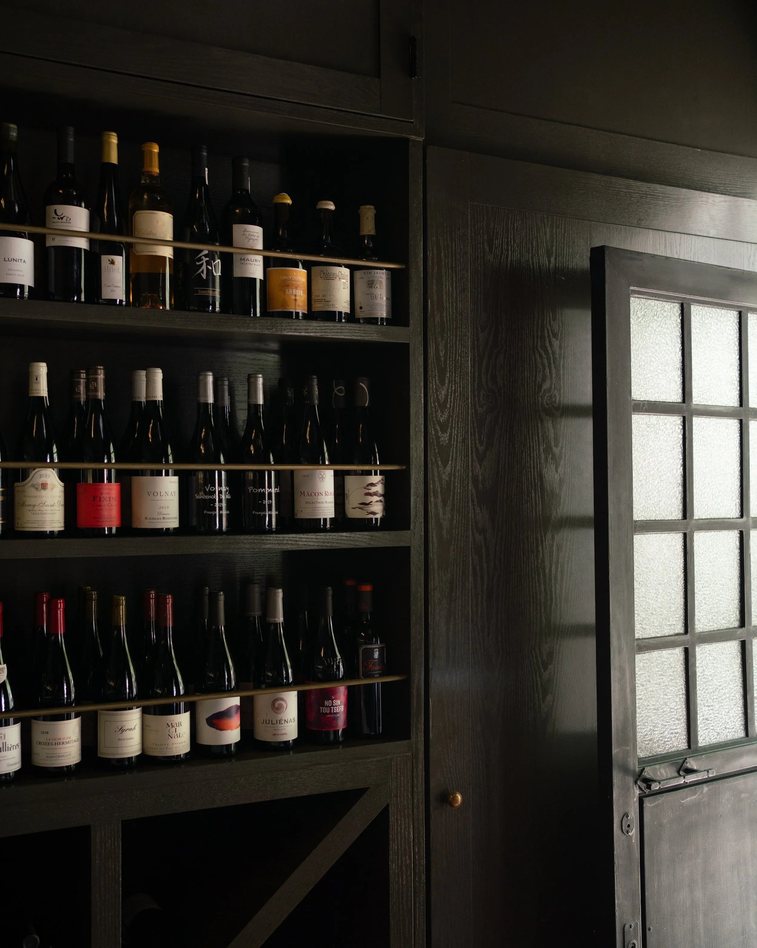 A dark wood wine cabinet with shelves filled with wine bottles, next to a door with glass panels.