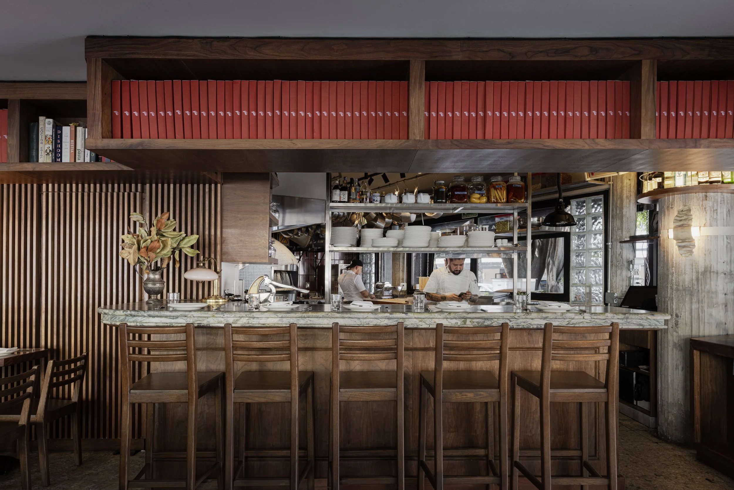 Interior of a modern restaurant with a bar counter, wooden chairs, and shelving with plates, spices, and kitchenware, with chefs working in the background.