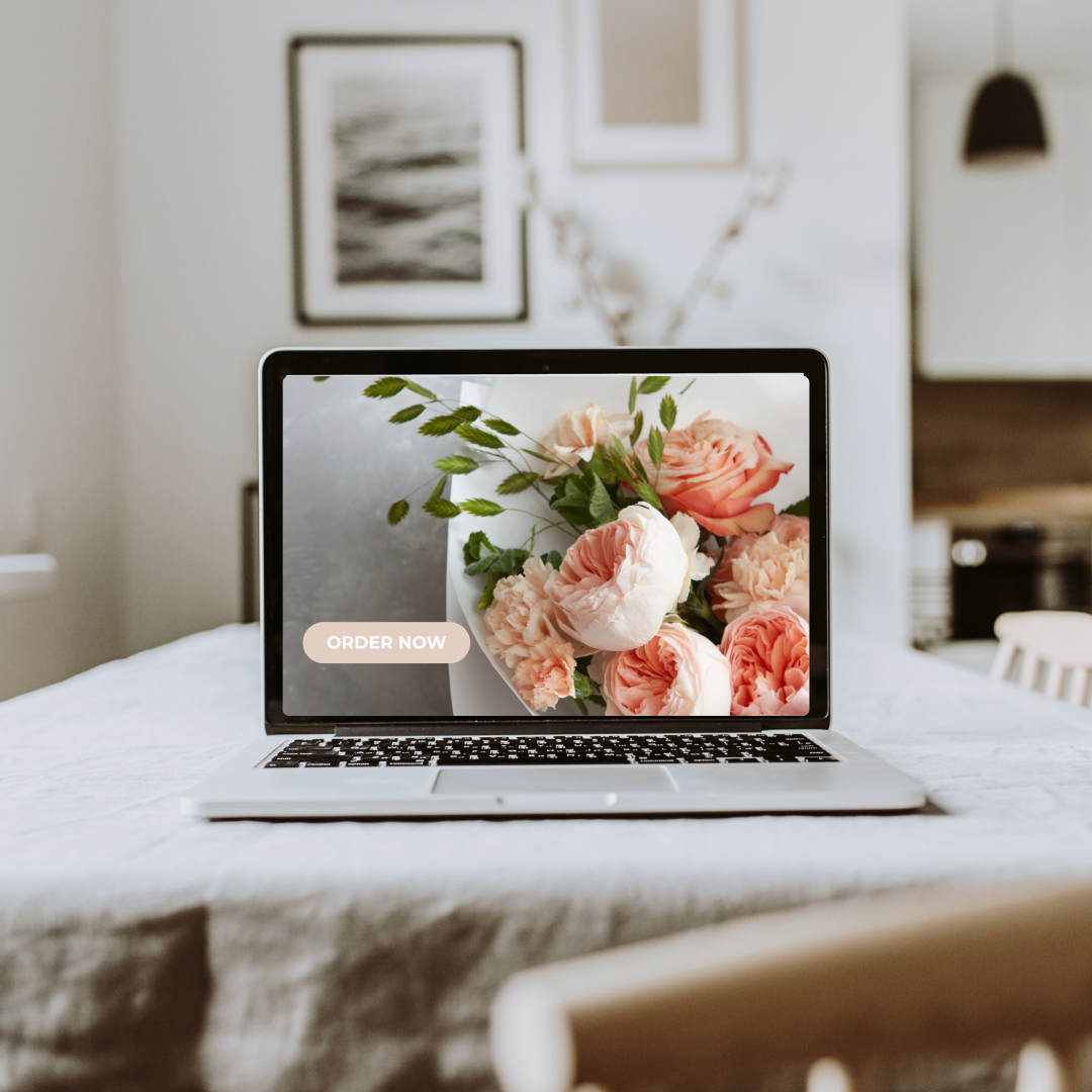 Laptop on a white table showing a bouquet of pink and white roses with a "Order Now" button, in a modern room with framed pictures on the wall created by Clair Creative branding and web design in Newmarket, Huntsville, Ottawa, Kawartha Lakes