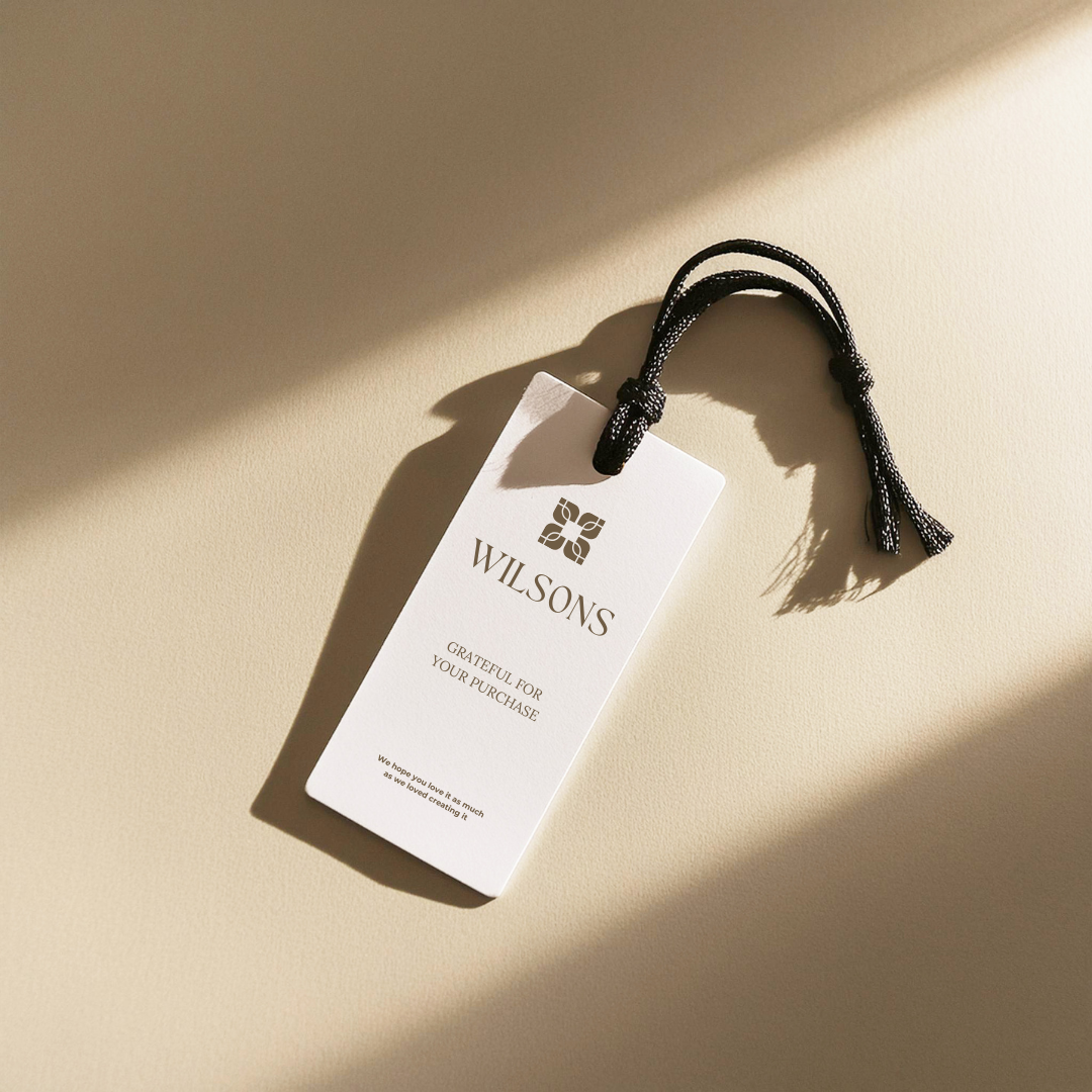 A white shopping or gift tag with the text "WILSONS GRATFUL FOR YOUR PURCHASE" and a black string, casting a shadow on a light beige surface with sunlight created by Clair Creative branding and web design in Newmarket, Barrie, Orillia, Collingwood,