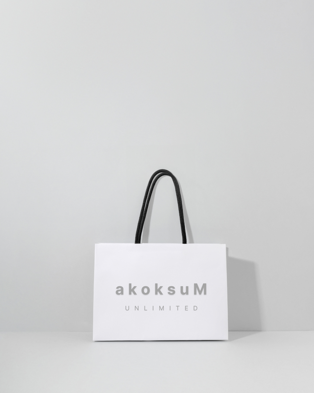 White shopping bag with black handles and the text 'akoksuM UNLIMITED' on it, placed against a plain light gray background. The brand was created by Clair Creative Branding and Web Design in Newmarket, Barrie, Collingwood, Muskoka