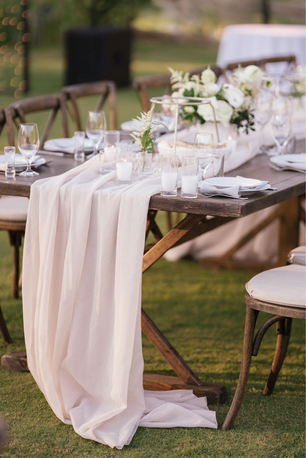 An outdoor wedding or event table decorated with white flowers, candles, and a long white table runner, set on a grassy lawn with wooden chairs. Outdoor wedding vs indoor wedding?
