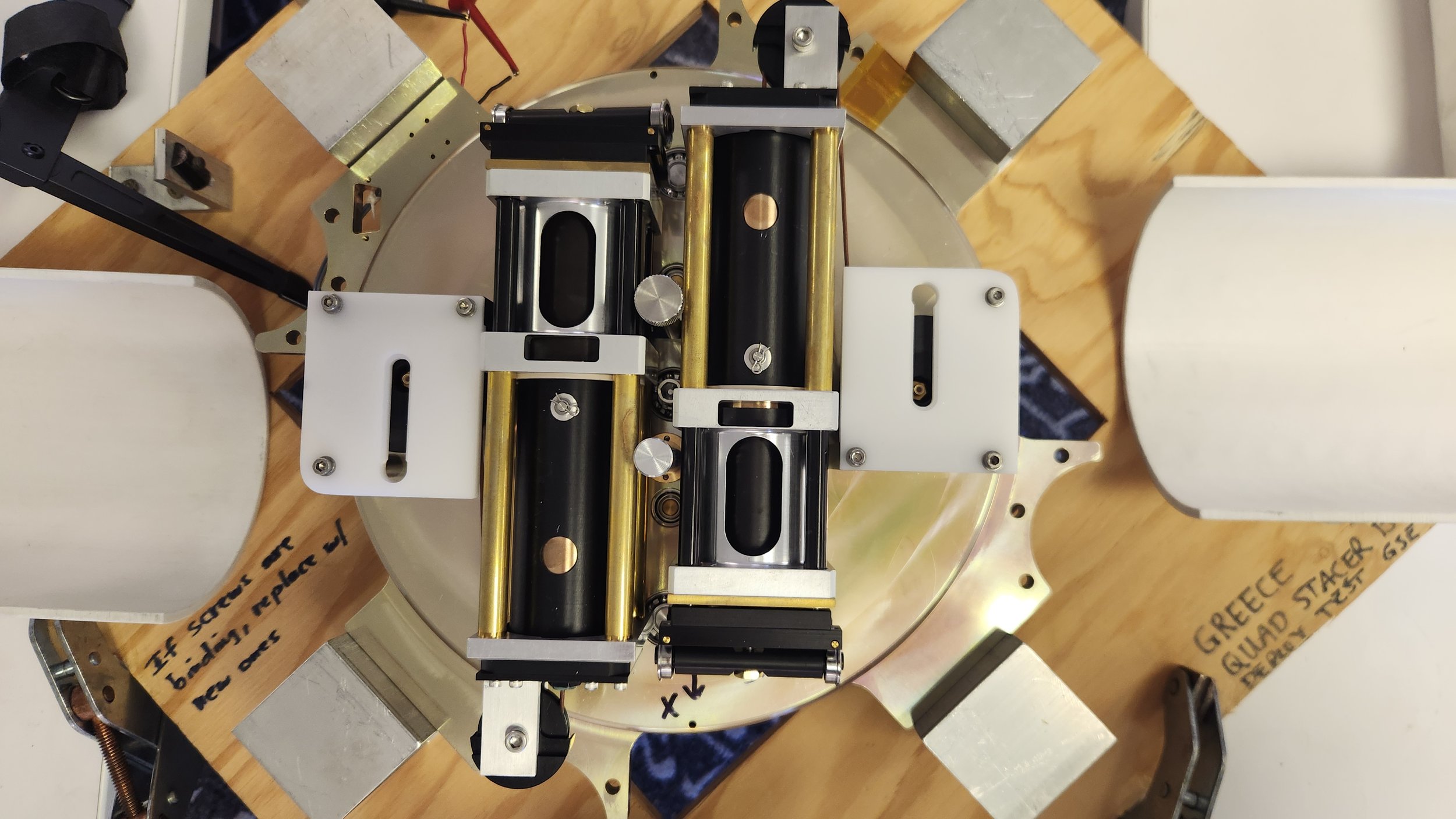 A scientific or mechanical apparatus placed on a wooden surface, with various black, gold, and white components, metallic parts, and screws.