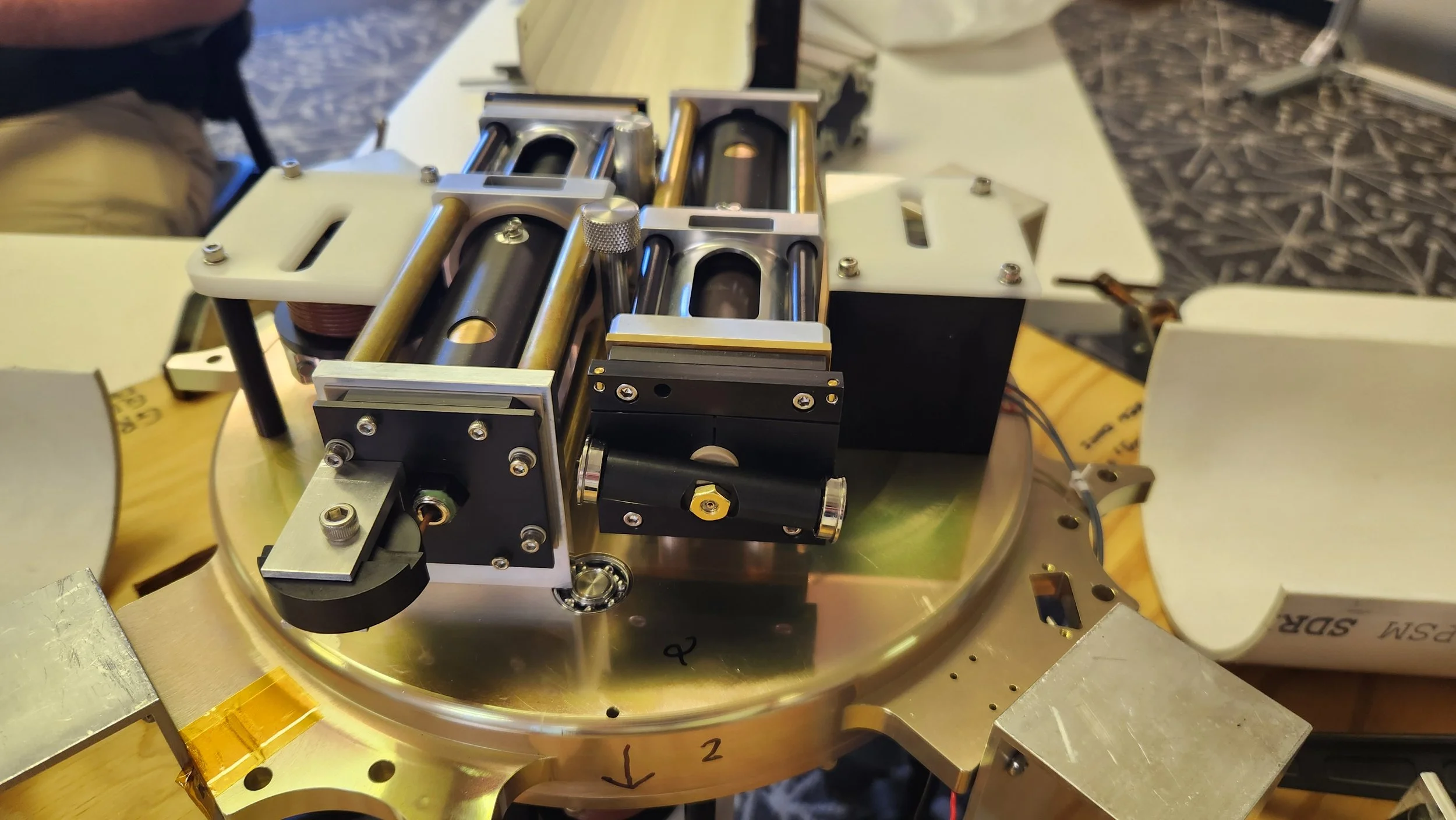 Close-up of a mechanical device with metal and black components, mounted on a circular metal base, with various rods, screws, and adjustable parts, sitting on a wooden surface.