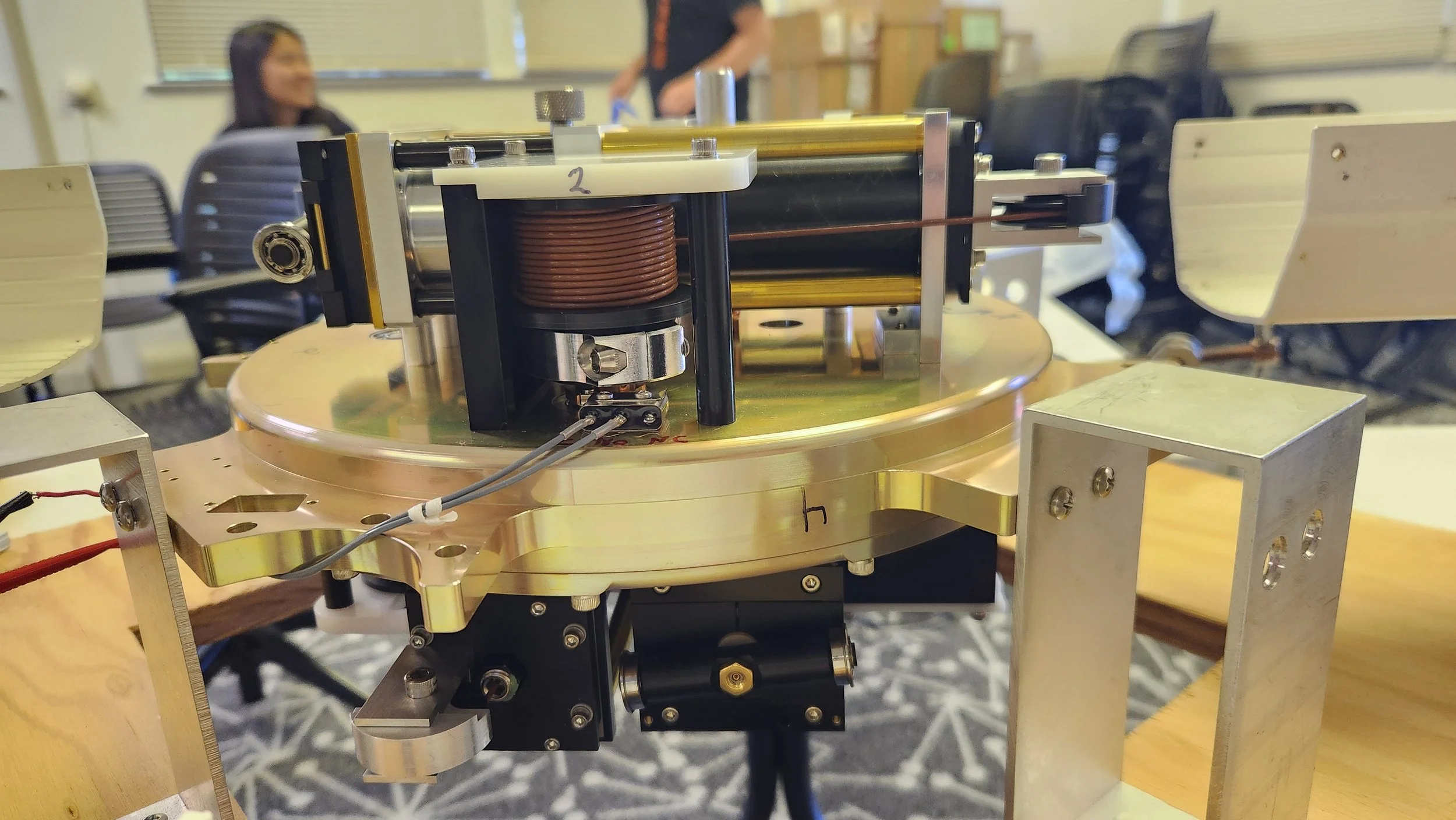 Close-up of a scientific or mechanical device with metallic components and copper coils, situated on a round metallic platform, in a room with chairs and people in the background.