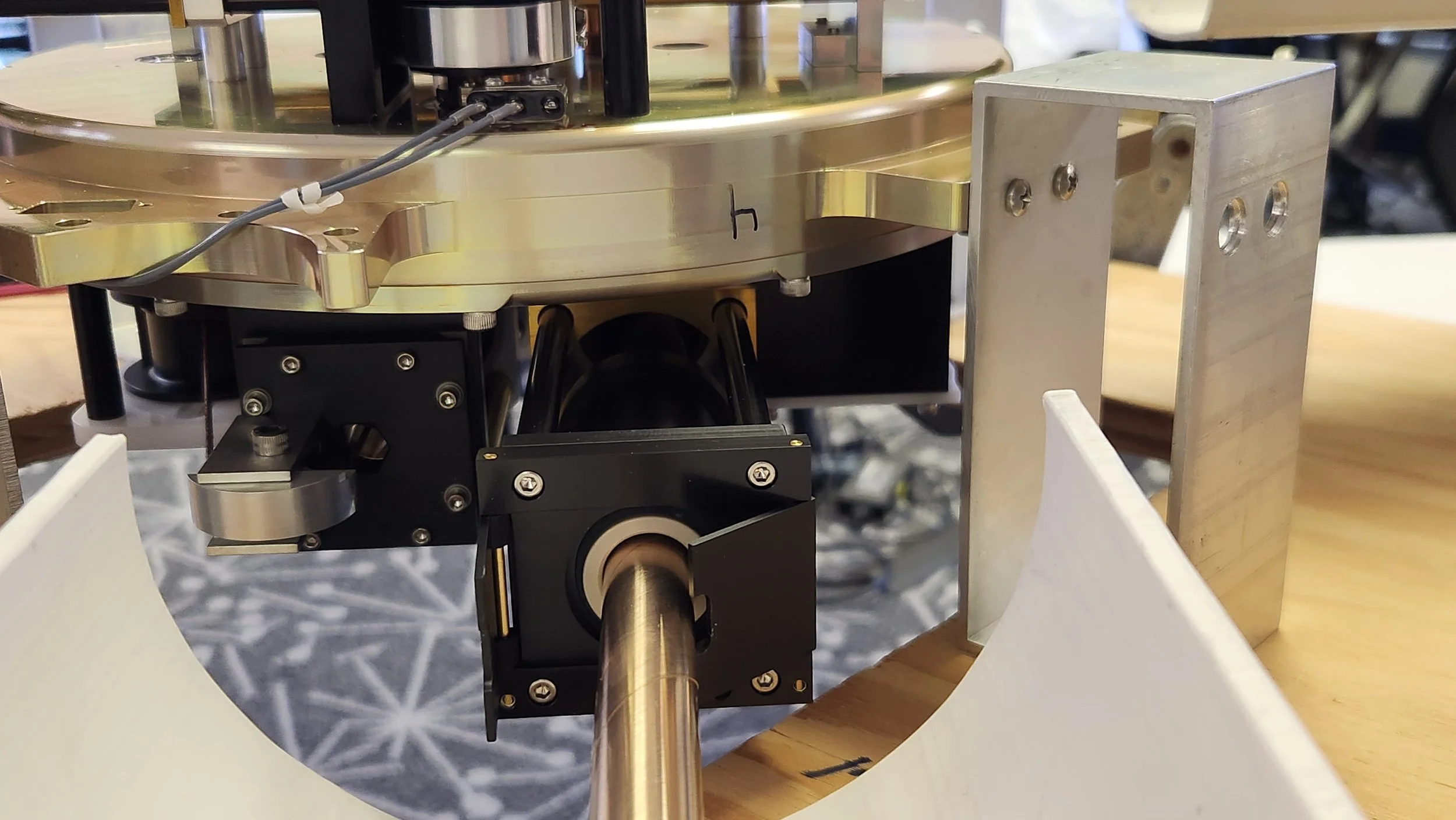 Close-up of a mechanical device with a metallic circular base, a black mounting bracket, and a long metal shaft. The device is assembled on a light-colored wooden surface with some structure and cables, possibly part of a scientific or industrial equ