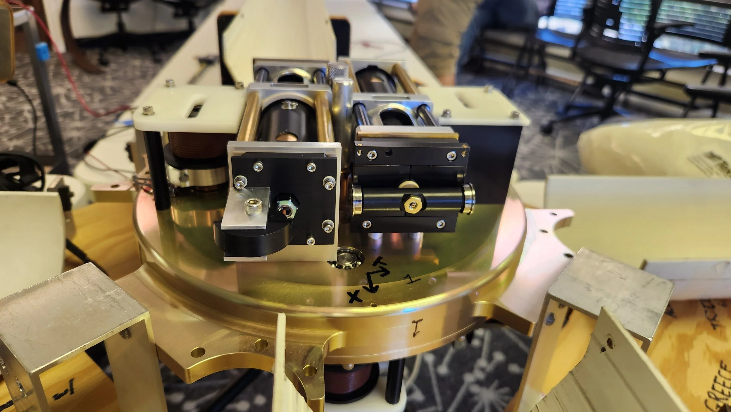 Close-up of a scientific or engineering apparatus mounted on a round, metallic base with electrical components, screws, and mechanical parts, set on a work surface with chairs and equipment in the background.