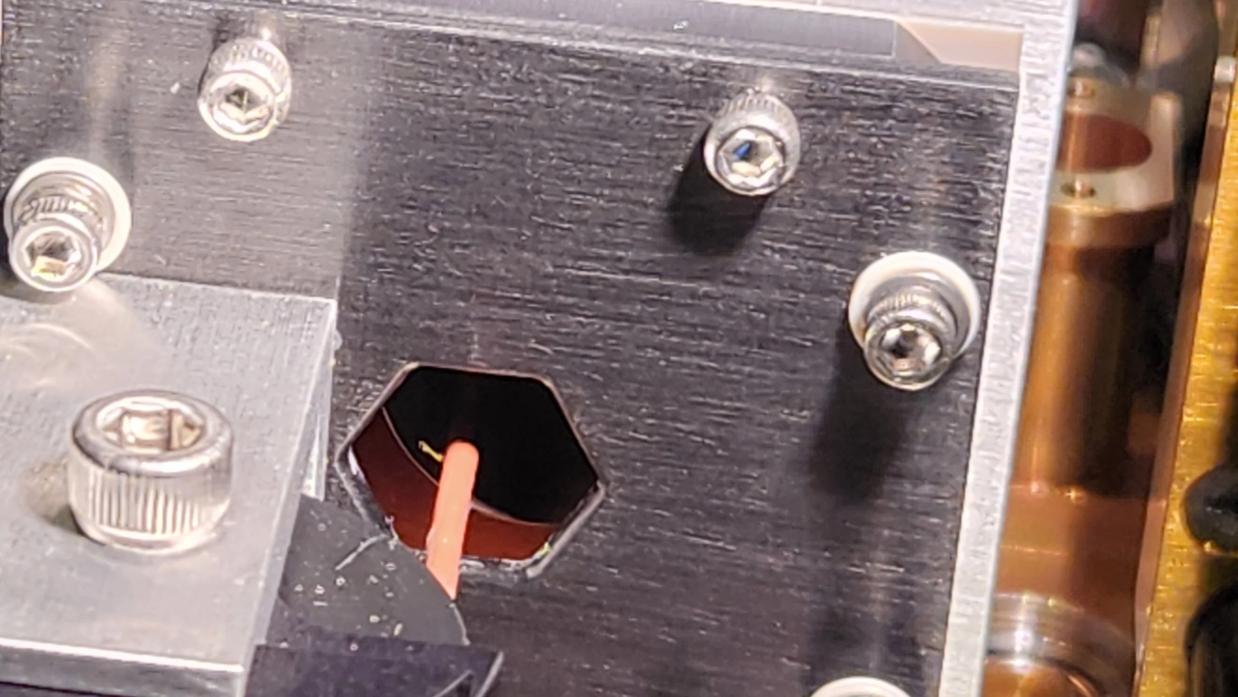 Close-up of a metal electronic component with screws, an orange wire connecting through a hexagonal hole, and some metallic and wooden parts in the background.