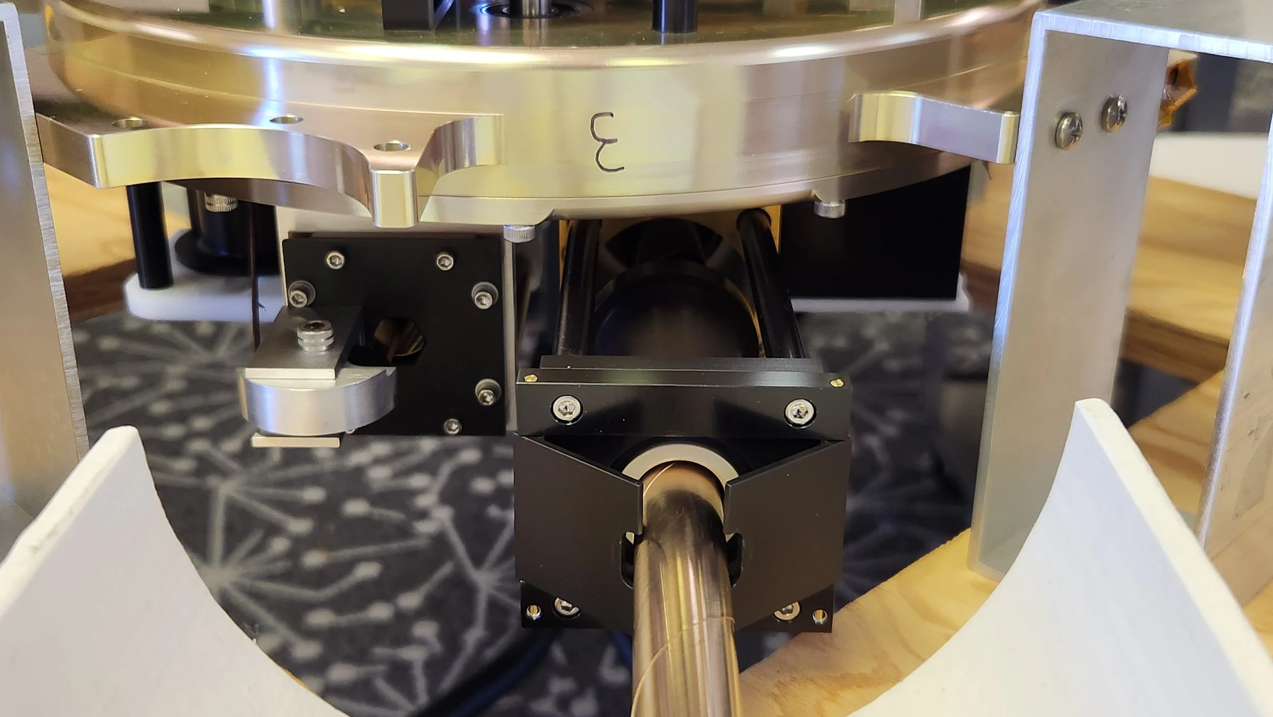 Close-up of a scientific or industrial apparatus with metallic components, black mounts, screws, and a long metallic rod, placed on a wooden surface and partially surrounded by white triangular panels.