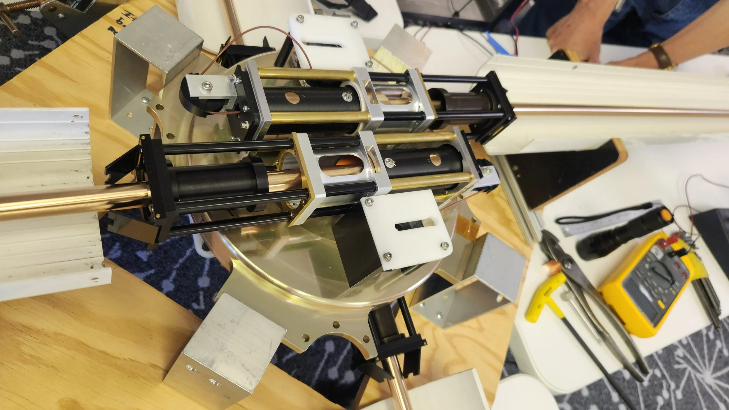 A robotic or mechanical device with metal and black components on a wooden table, surrounded by tools like a multimeter, screwdriver, and a yellow-handled tool, in a workshop or lab setting.